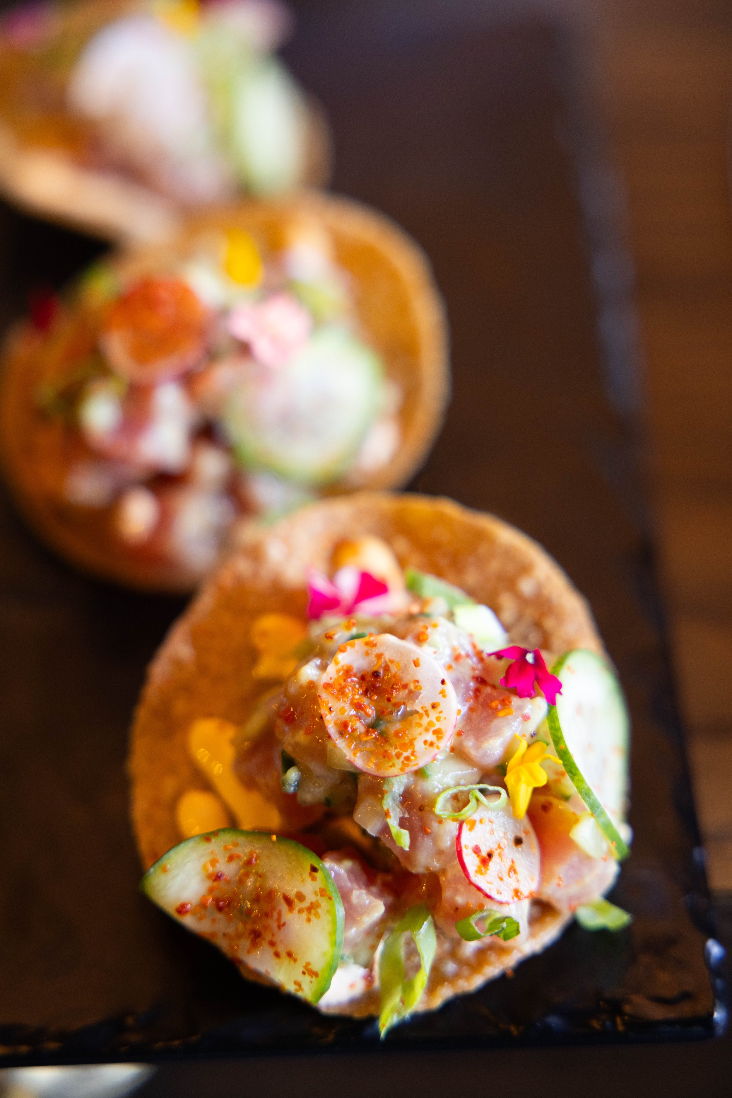 Close-up of two tacos on a black platter, filled with diced seafood, sliced radishes, cucumber slices, sprouts, and garnished with edible flowers.
