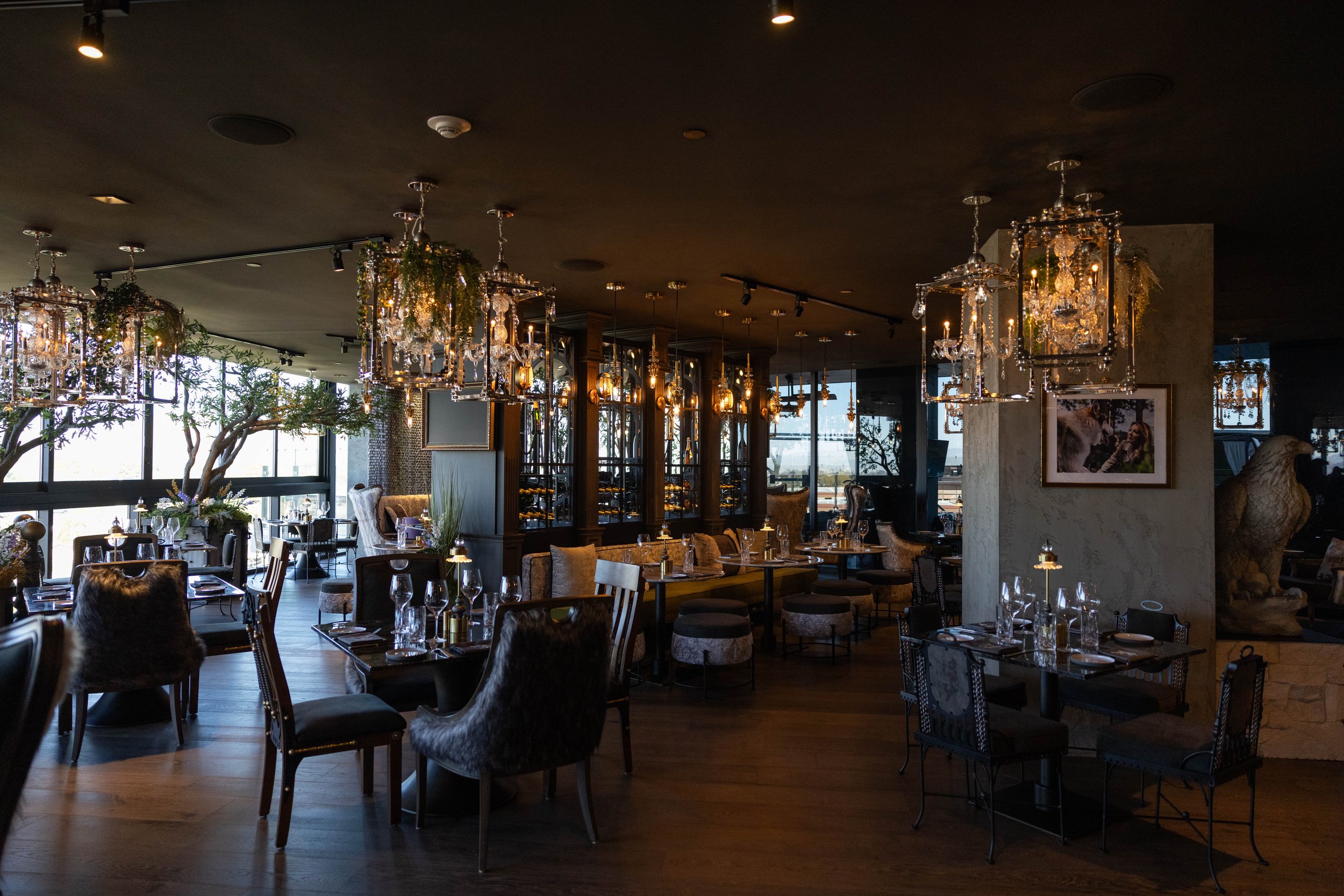 Elegant restaurant interior with chandeliers, wooden floors, and large windows, set up for dining with tables, chairs, and glassware.
