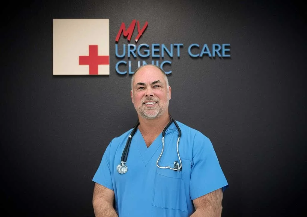 A male healthcare professional with a beard wearing blue scrubs and a stethoscope around his neck in a hospital setting.