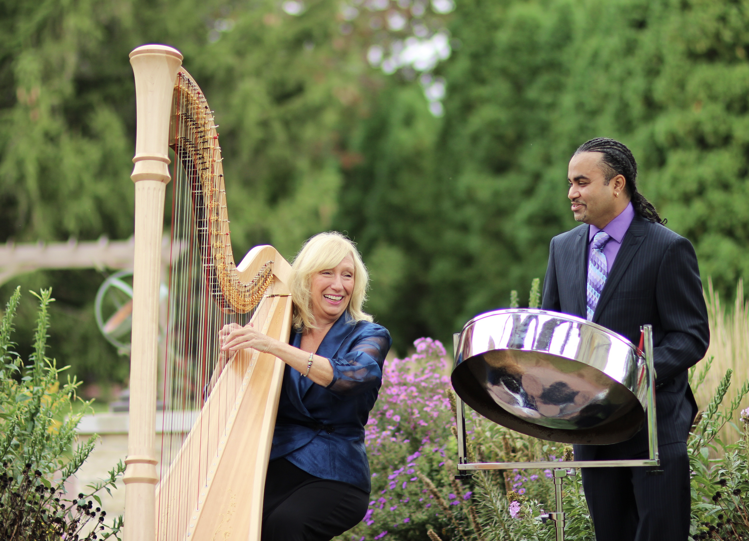 Two musicians, a woman and a man, playing musical instruments outdoors in a garden with green trees and purple flowers. The woman is playing a harp, and the man is playing steel drums.