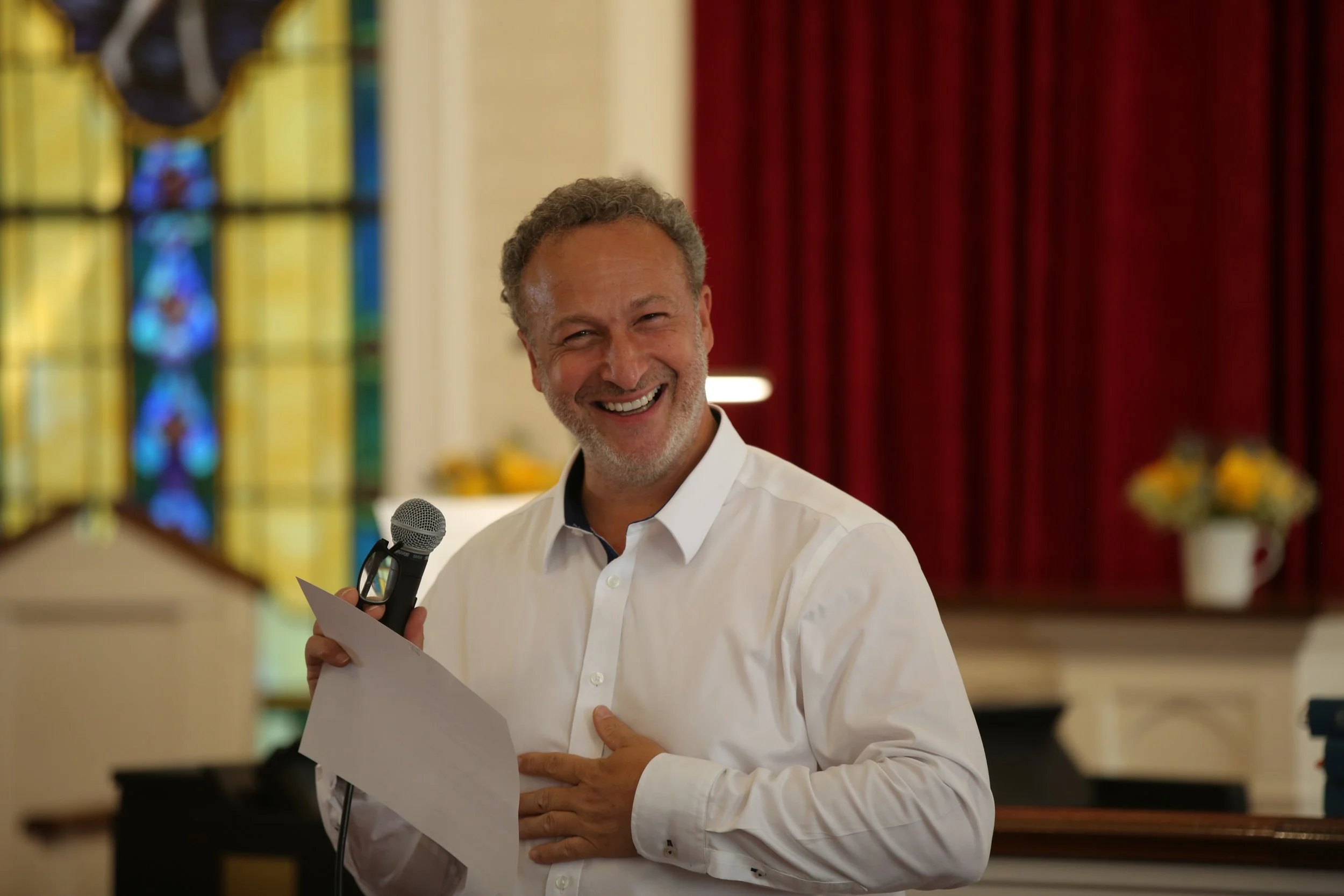 A man with gray curly hair and a beard, smiling, wearing a white dress shirt, holding a microphone and papers, standing indoors with stained glass windows and red curtains in the background.