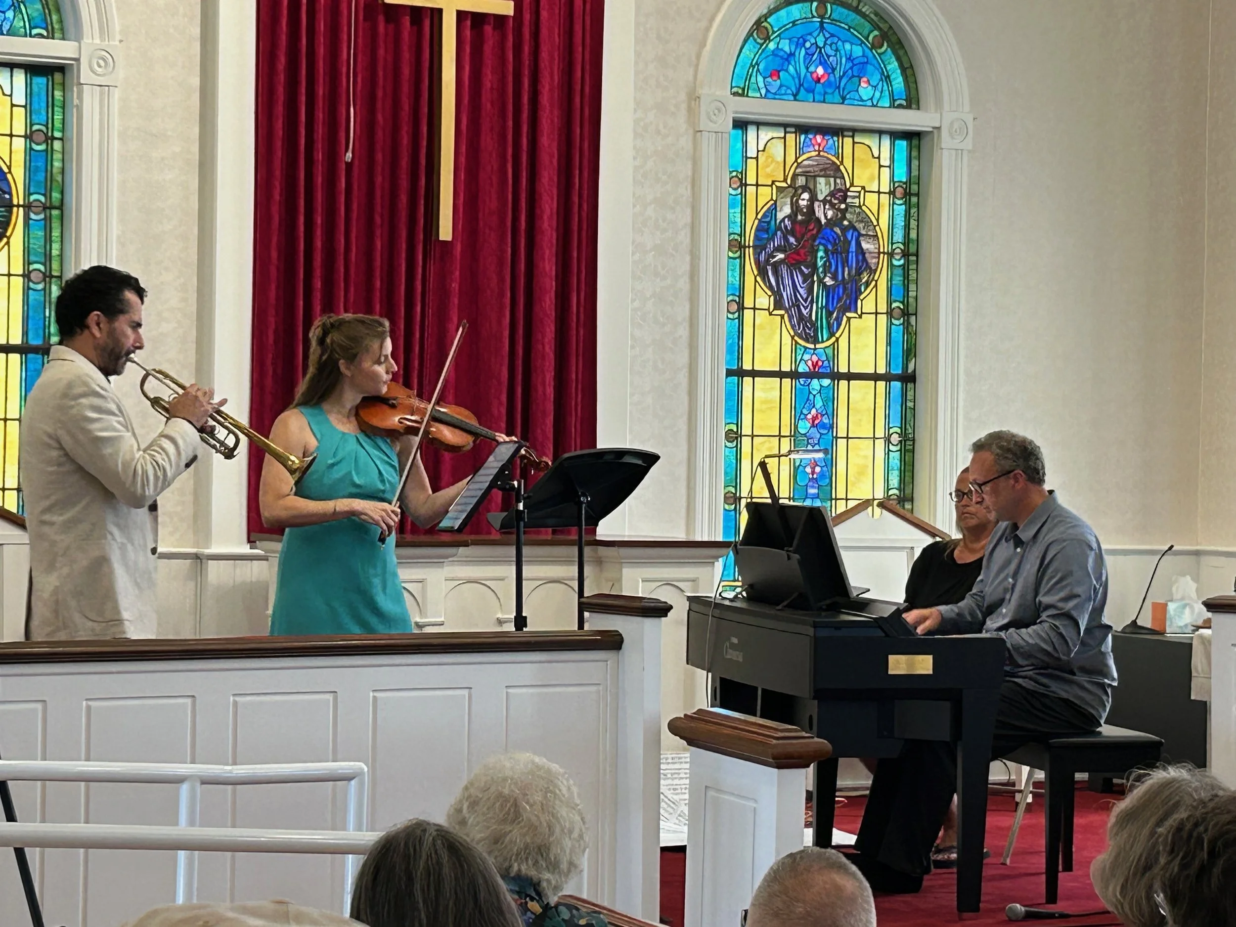 Musicians performing a religious ceremony in a church, with stained glass windows behind them. One person is playing the trumpet, another is playing the violin, and a third is playing a keyboard piano.