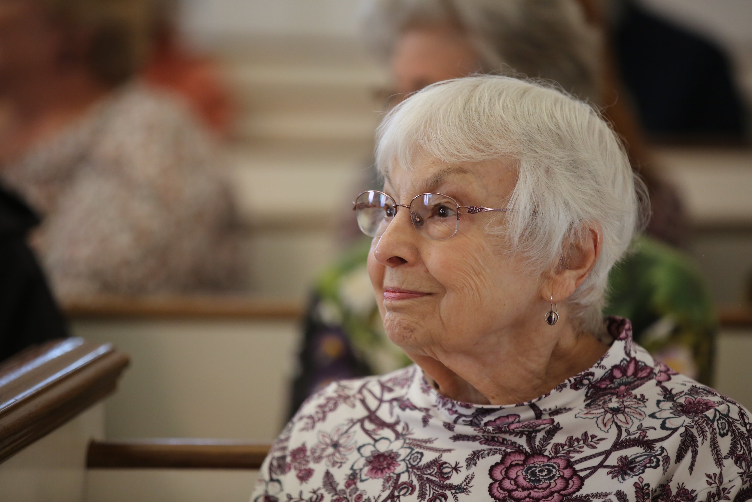 A senior woman with white hair and glasses talking to a man with glasses and gray hair. The woman is wearing a floral blouse and earrings. They appear to be in a social or community setting.