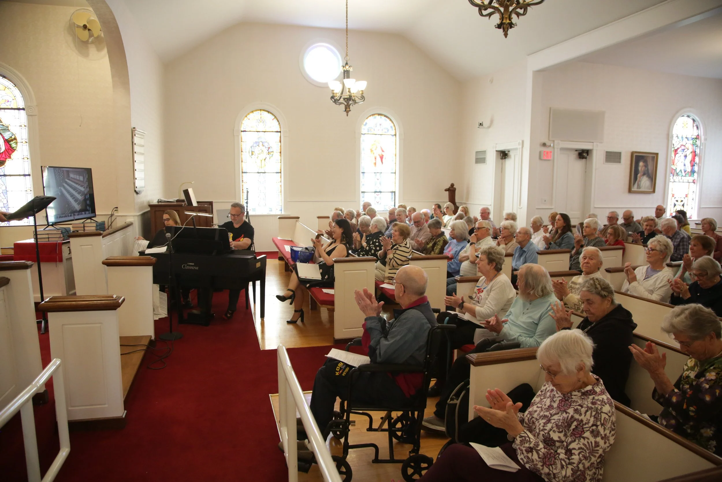 A congregation of people seated in a church, with stained glass windows, a chandelier, and a grand piano in the front.