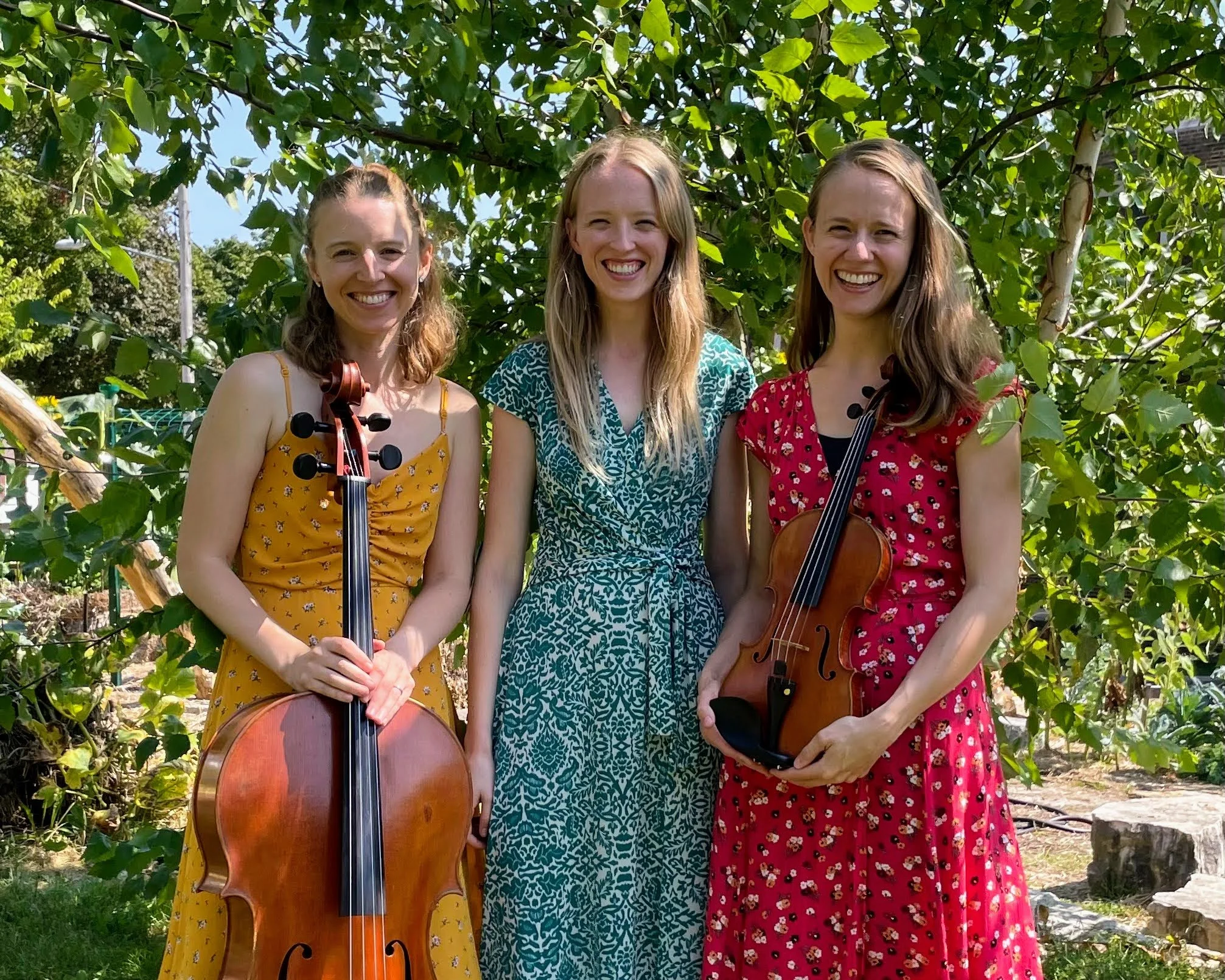 Three women smiling outdoors among green trees, two holding violins.