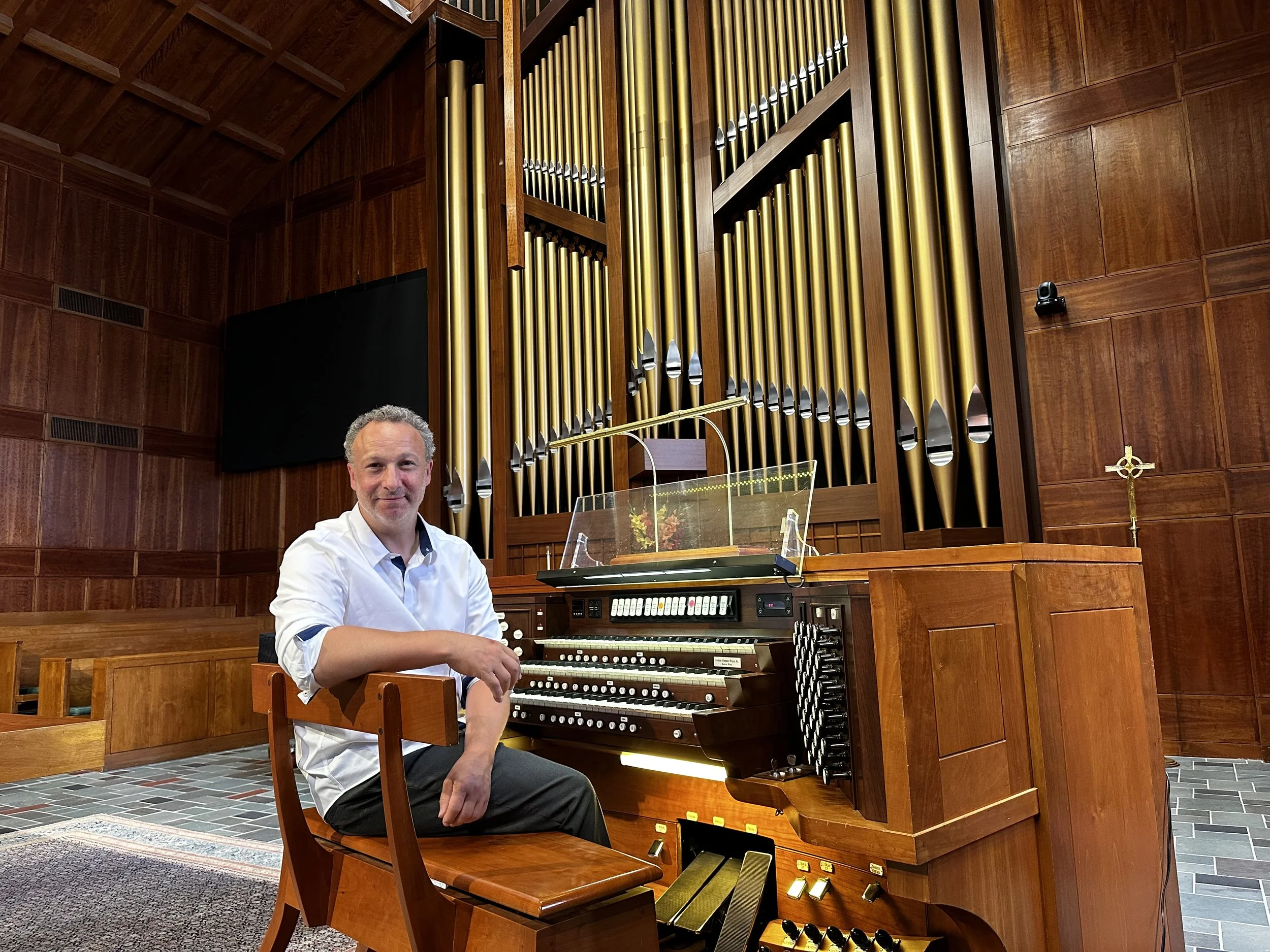A man in a white shirt sitting at a pipe organ inside a church or chapel, with wooden paneling and pipes in the background.