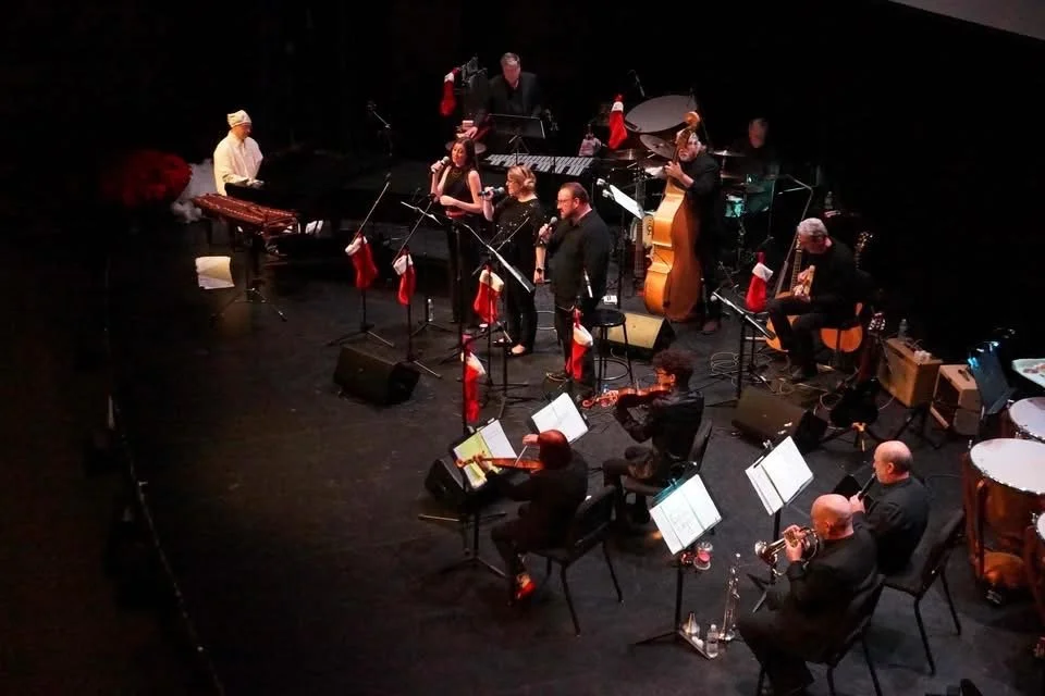 Musicians on stage performing with various instruments, including a grand piano, guitar, drums, double bass, and wind instruments, decorated with Christmas stockings.