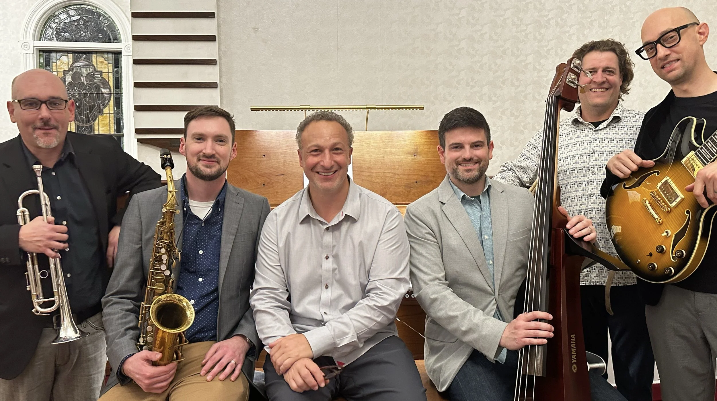 Six men are posing together indoors, each holding or standing near musical instruments, in front of a wooden piano and white wall with decorative wood slats and a stained glass window.