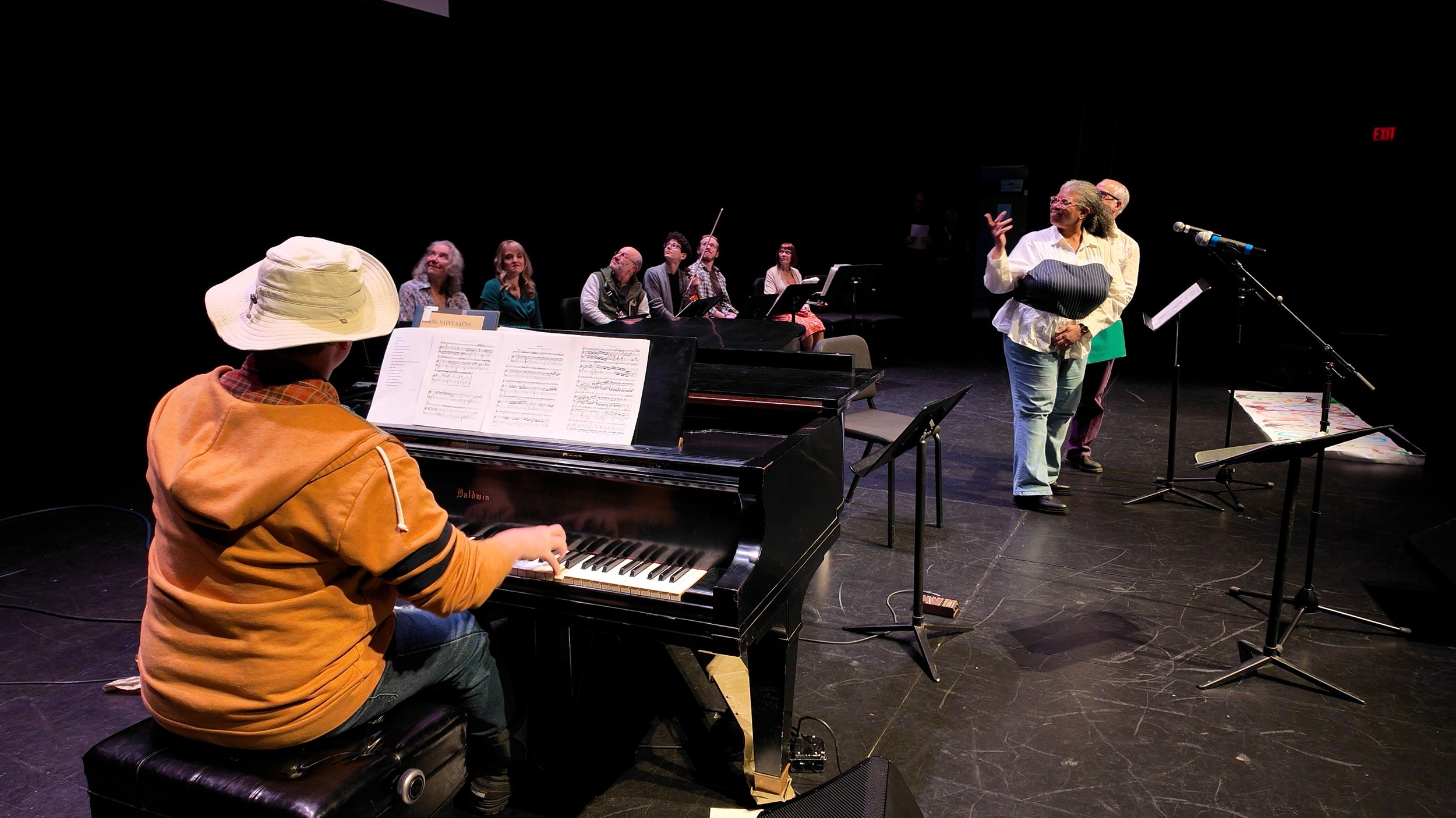 Music rehearsal on stage with a pianist, a choir of seven members, and a woman conducting or speaking at a microphone.