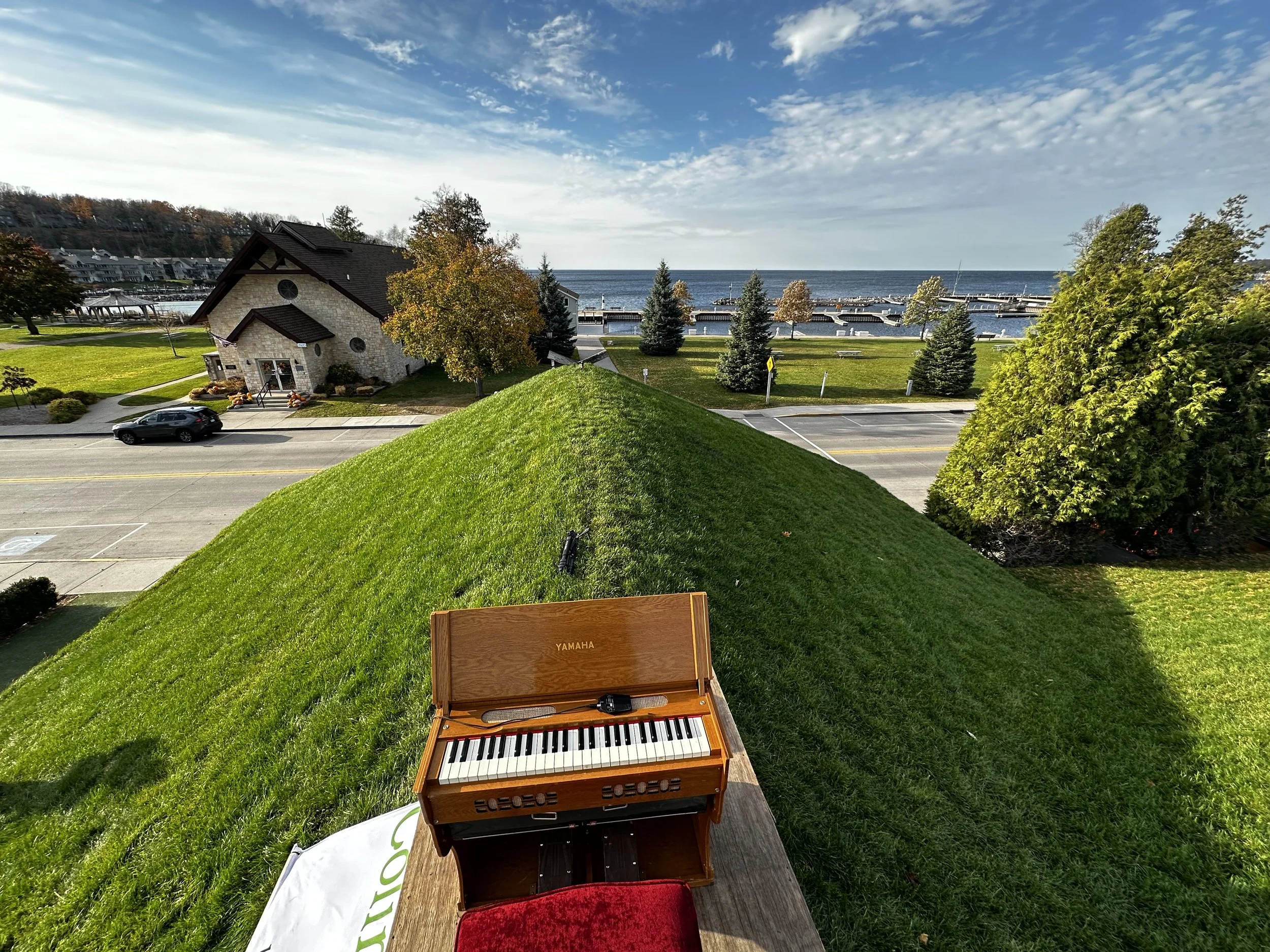 A view of a grassy area with a small wooden keyboard instrument in the foreground on a sunny day, overlooking a parking lot, trees, a building, and a body of water with a pier in the background.