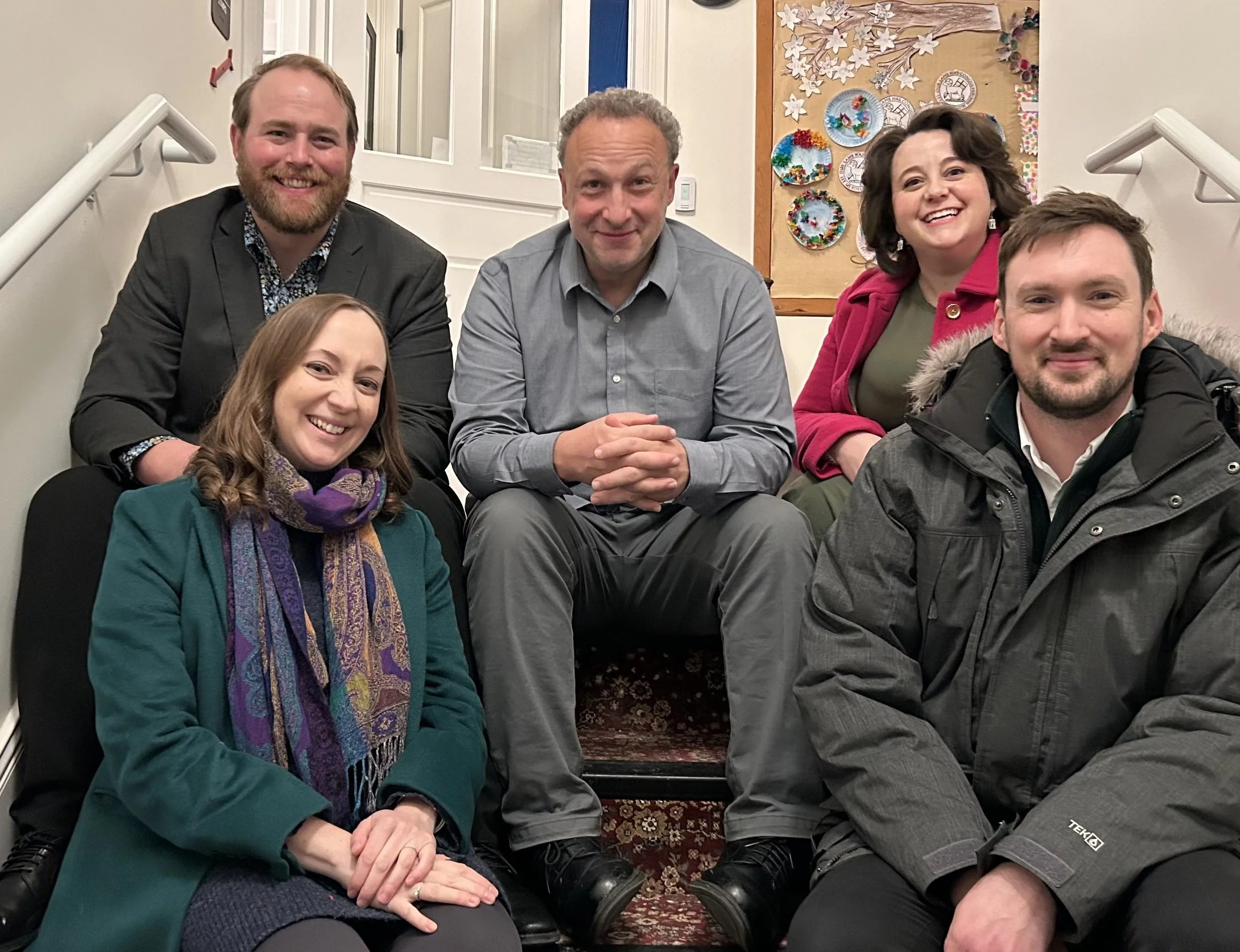 A group of five smiling people posing together in an indoor setting, with a colorful decorated bulletin board and stairs in the background.