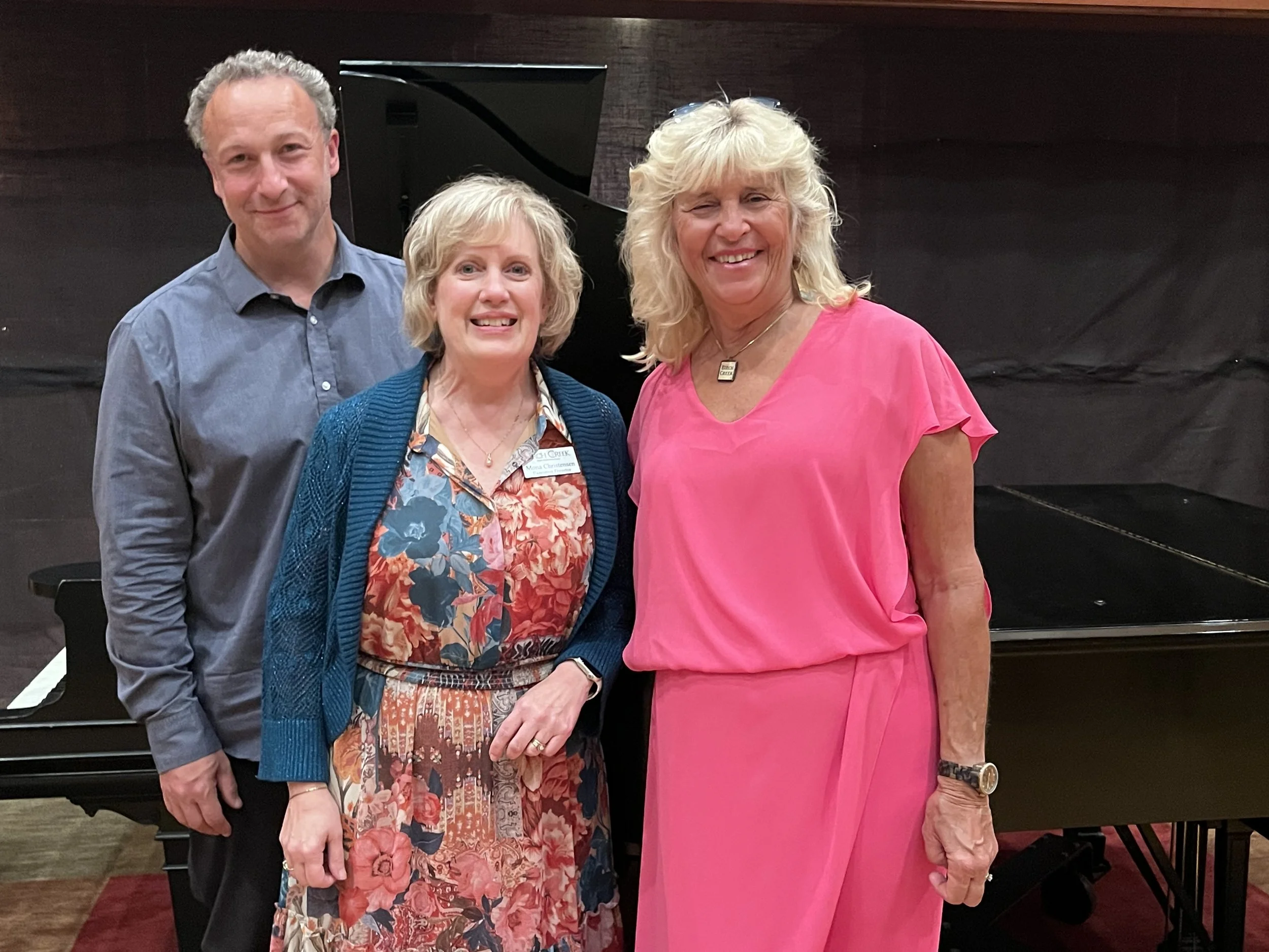 Four people, two women and two men, standing together indoors in front of a black piano, smiling for the photo.