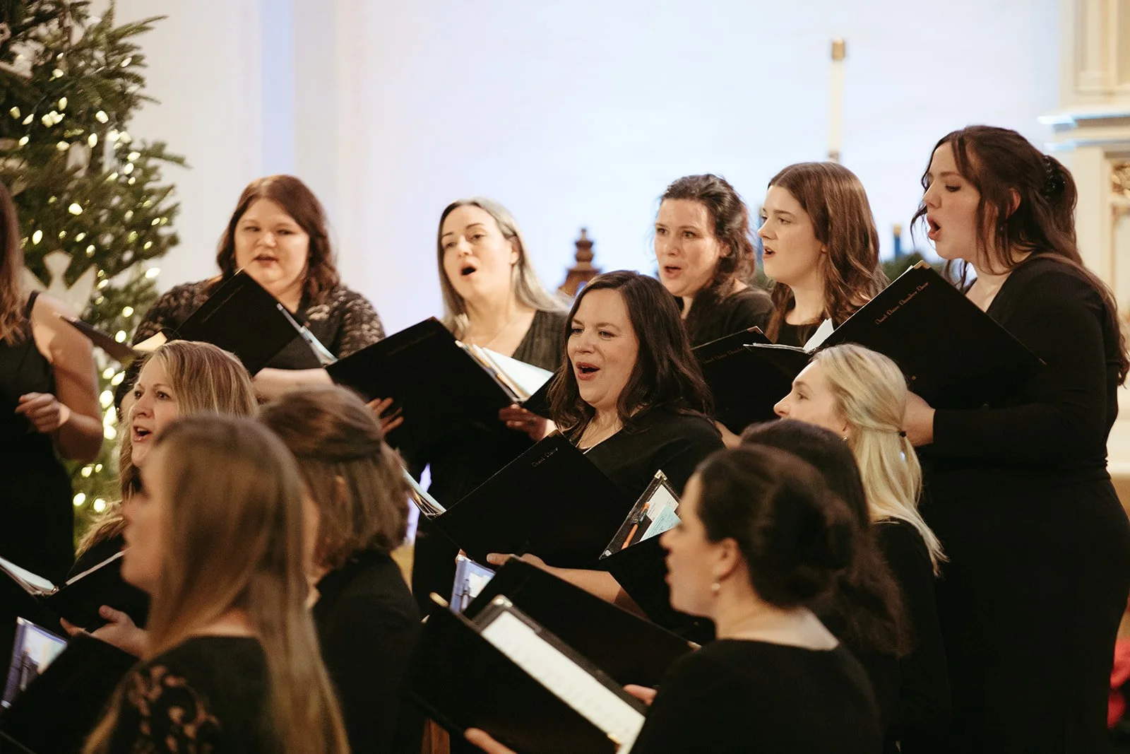 A group of women singing in a choir during a Christmas celebration, with a decorated Christmas tree in the background.