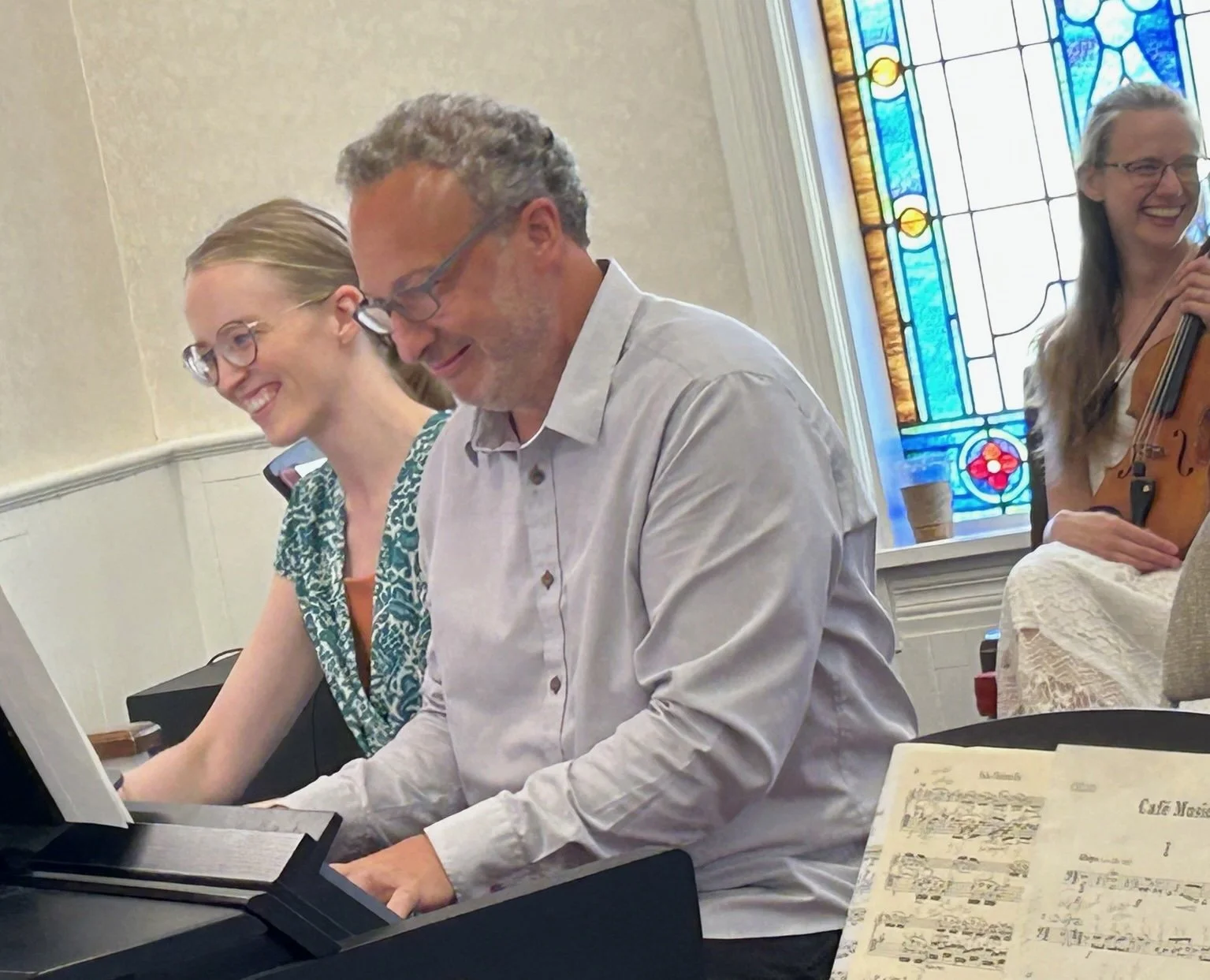 Three people smiling and playing musical instruments in a room with a stained glass window in the background. One person is playing a keyboard, another a violin, and the third is smiling while looking at sheet music on a stand.