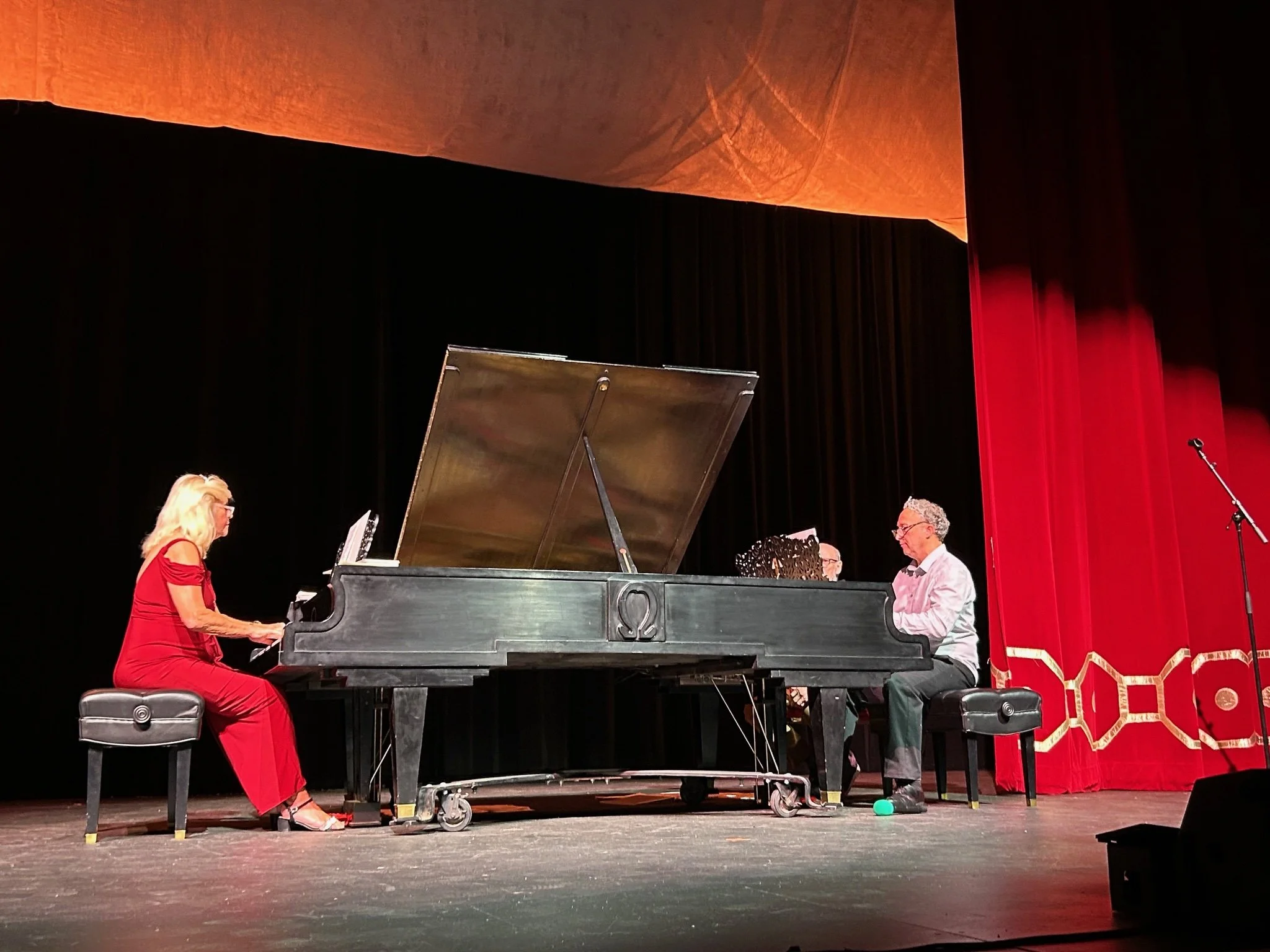 A woman in a red dress playing the piano on stage, with three men watching and listening, a grand piano, and a red curtain in the background.