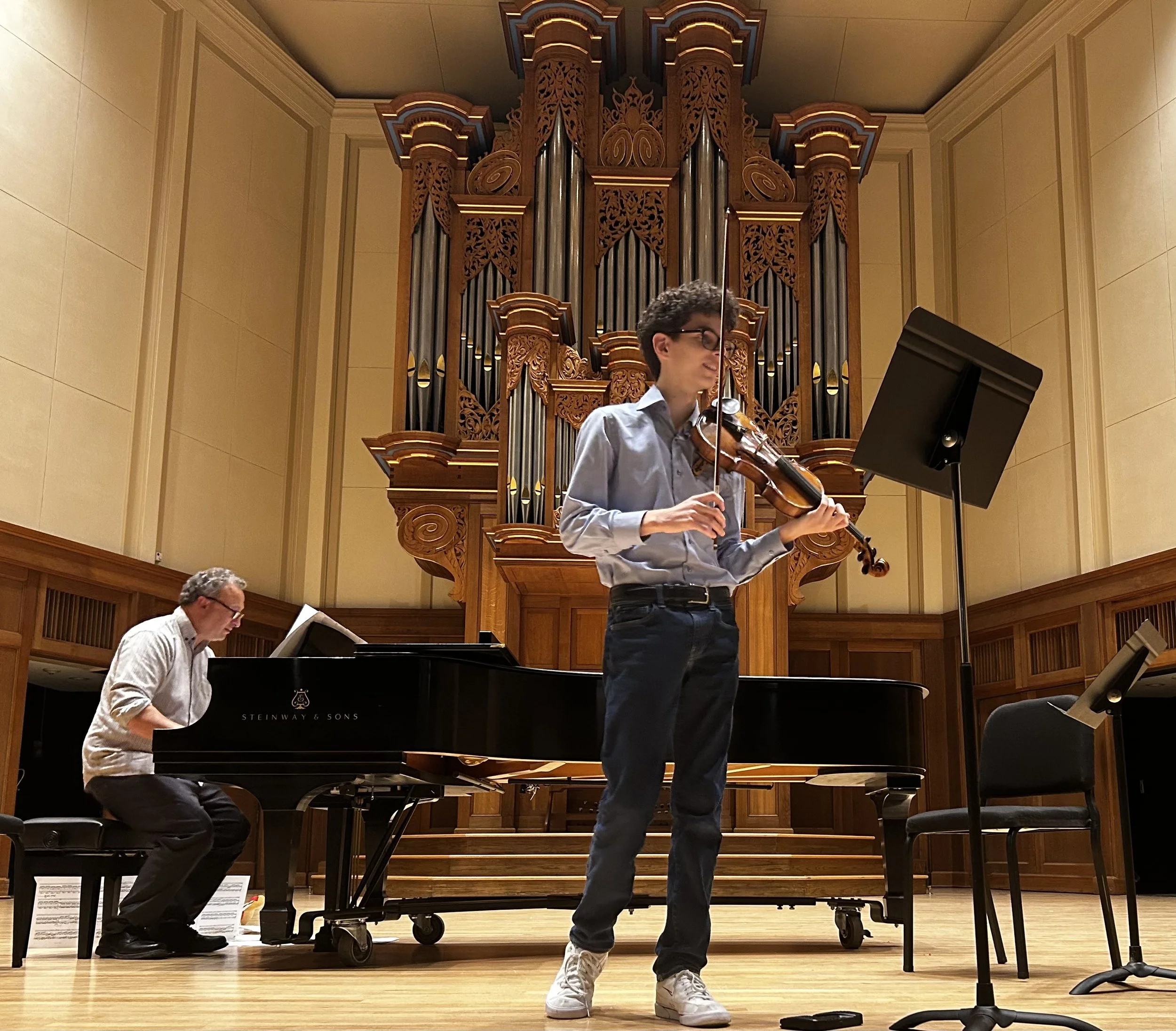 A musician playing the violin on stage with an organ in the background. The stage has wood paneling, and a pianist is seated at a black grand piano to the left.