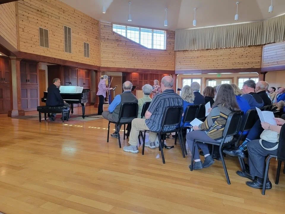 Group of people seated in chairs facing a stage with a woman speaking and a man playing piano in a wood-paneled room.