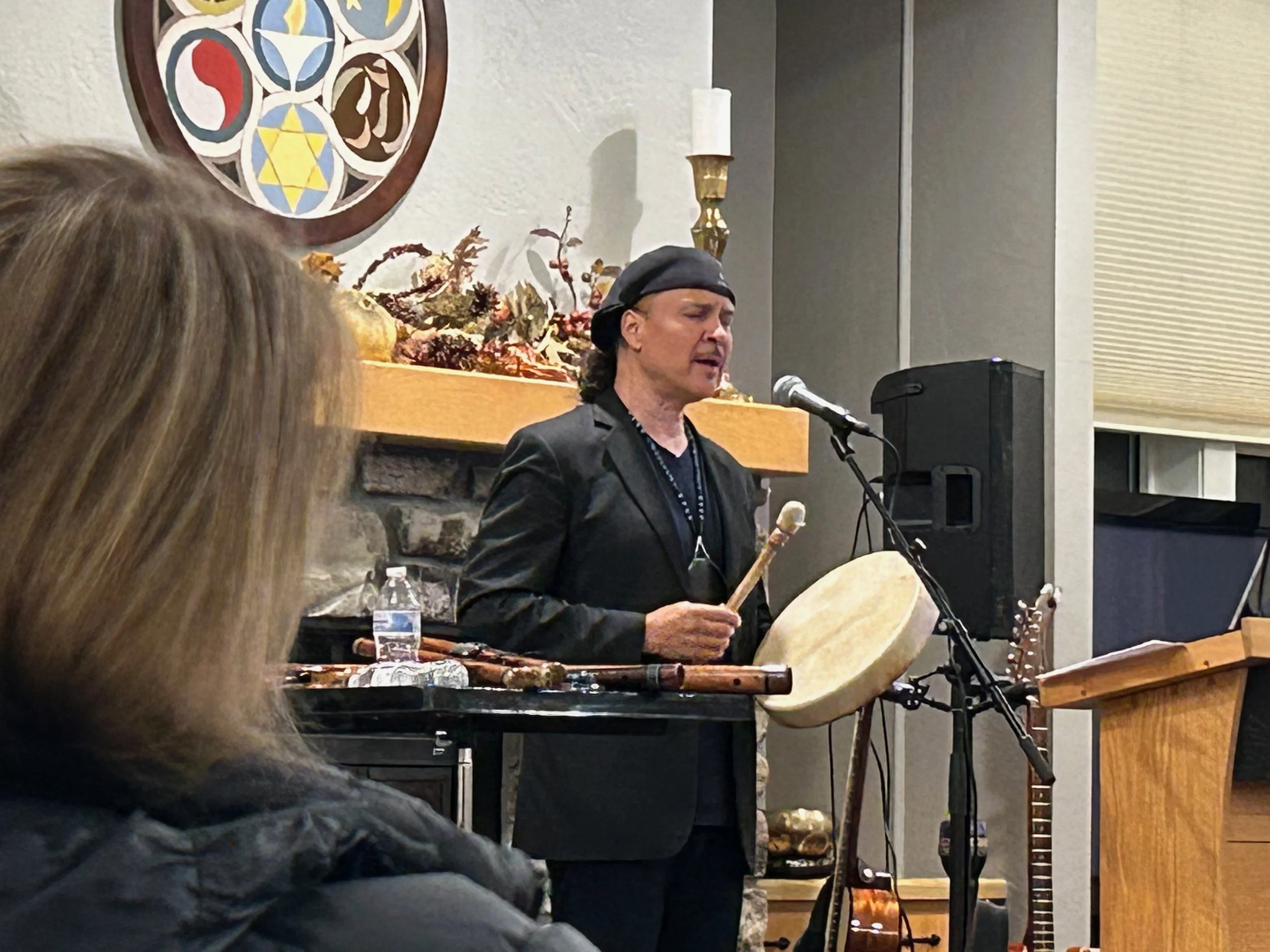 A man wearing a black hat and leather jacket is playing a drum and singing into a microphone. He is standing in front of a wall with religious or spiritual artwork, a candle, and decorative items. Part of an audience is visible in the foreground.