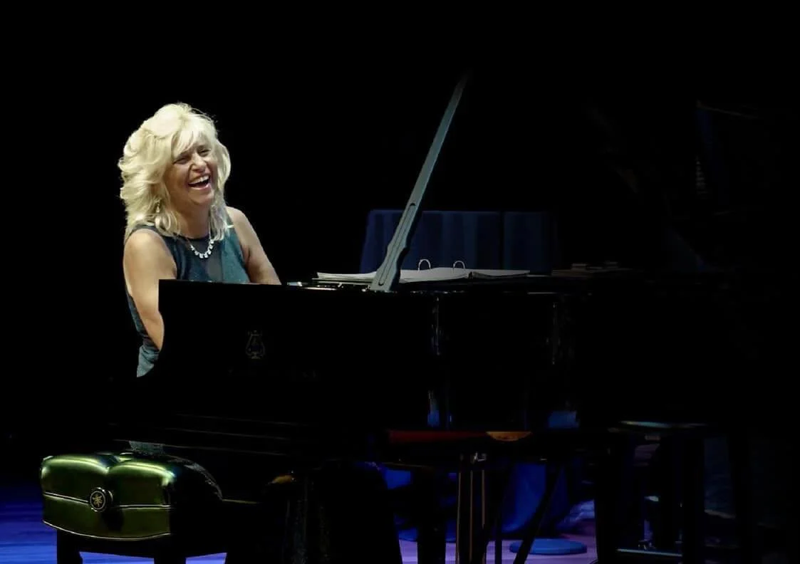 A woman with blonde curly hair laughing while sitting at a black grand piano on a dark stage.