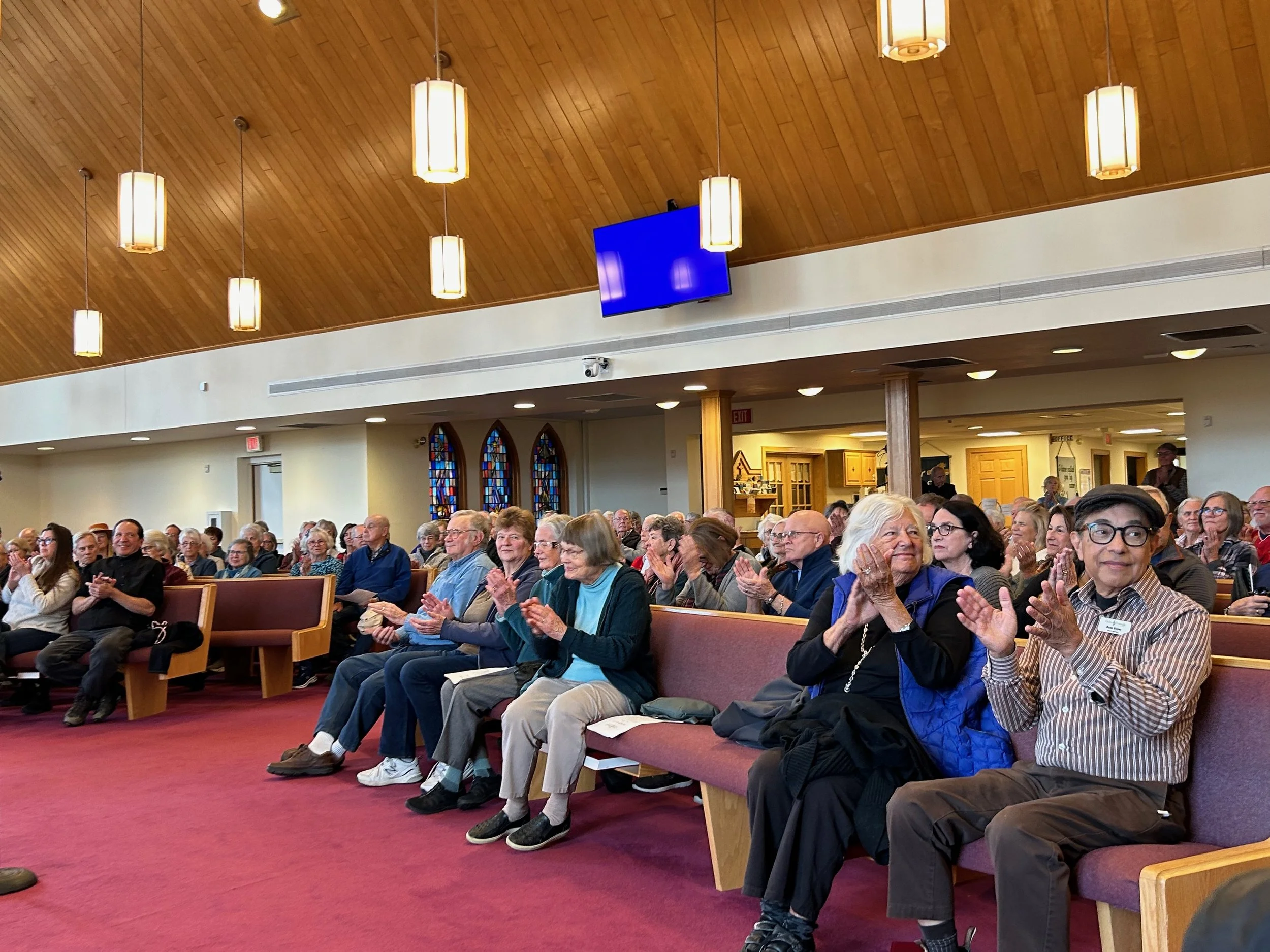 A large group of mostly elderly people seated in pews inside a church, attending a service or event, with some clapping.