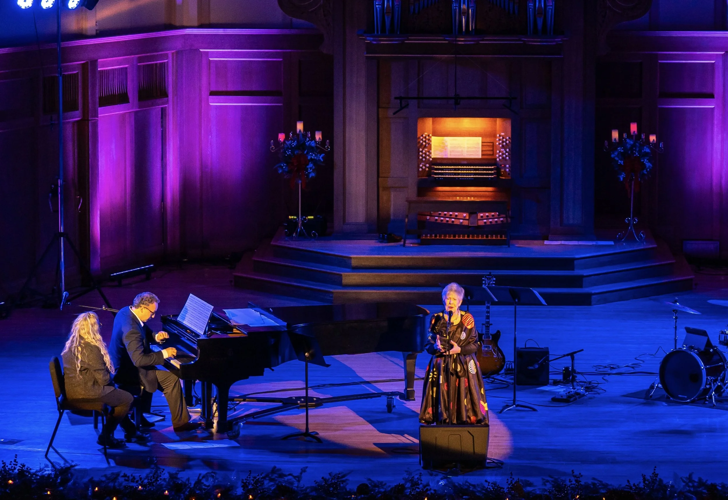 A musical performance on stage with a woman singing into a microphone, a pianist playing a grand piano, and a man playing a violin. The stage is decorated with purple lighting and Christmas floral arrangements.