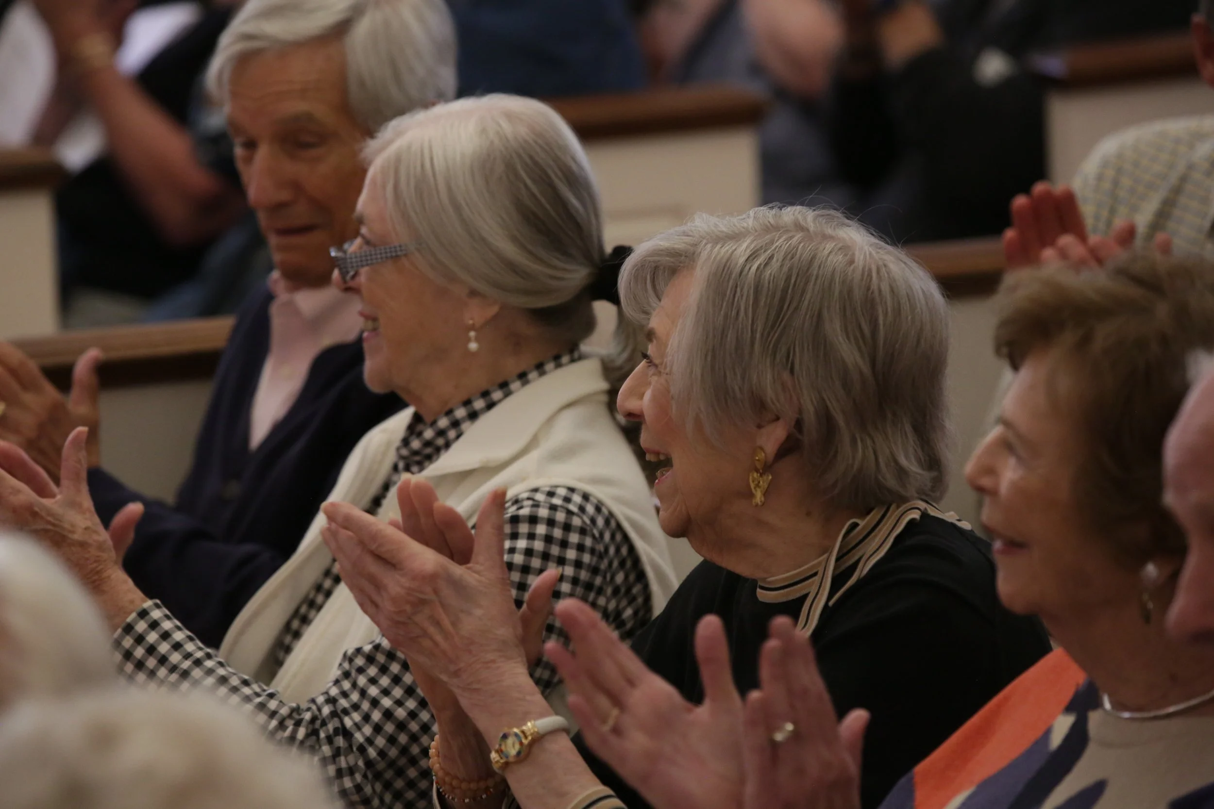 A group of elderly women sitting closely together in a church or auditorium, clapping and smiling, during an event or service.