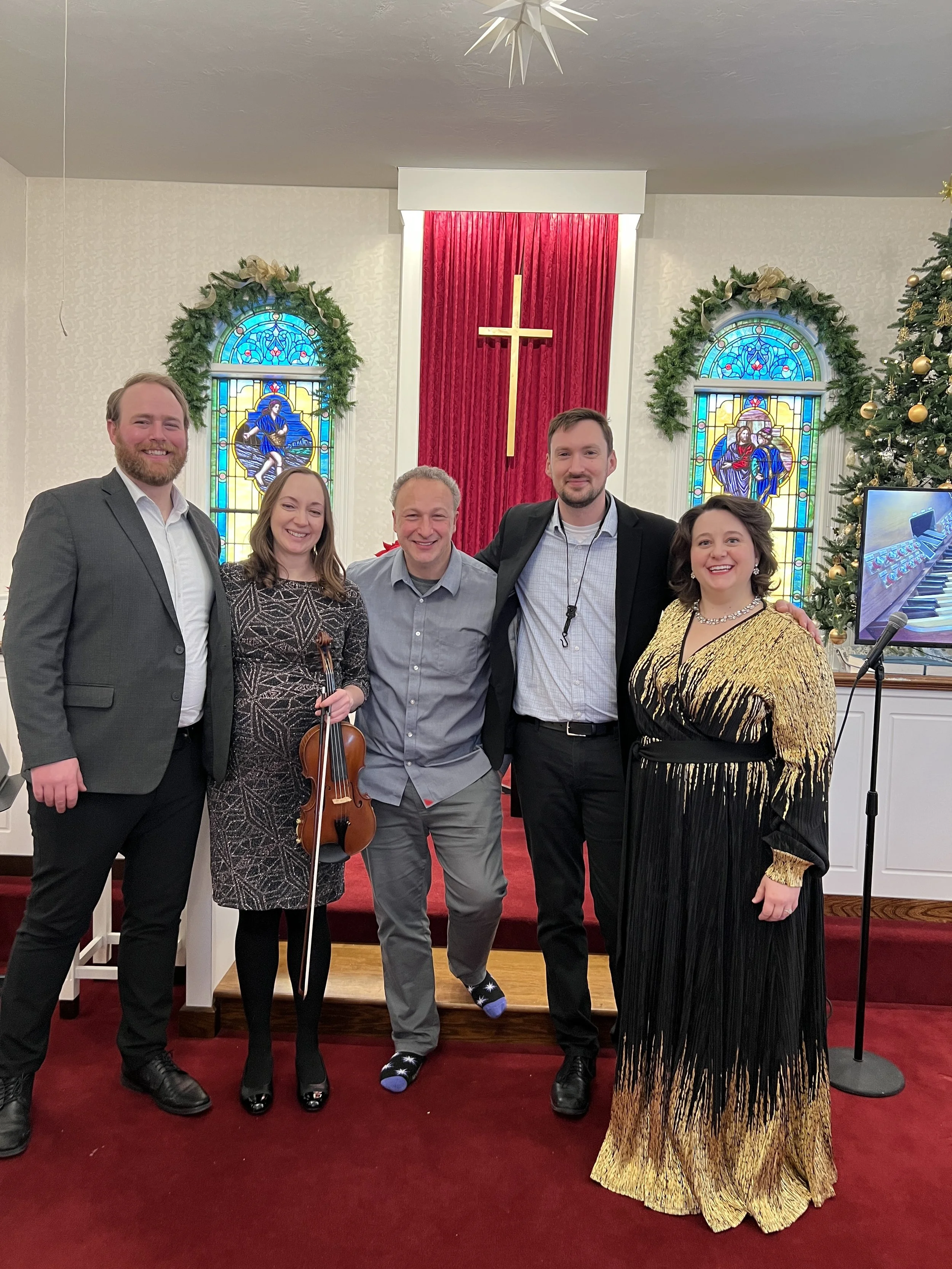 Group of five people standing inside a church with stained glass windows and a decorated Christmas tree. They are smiling and dressed in formal and festive attire, with a cross and red velvet curtain in the background.