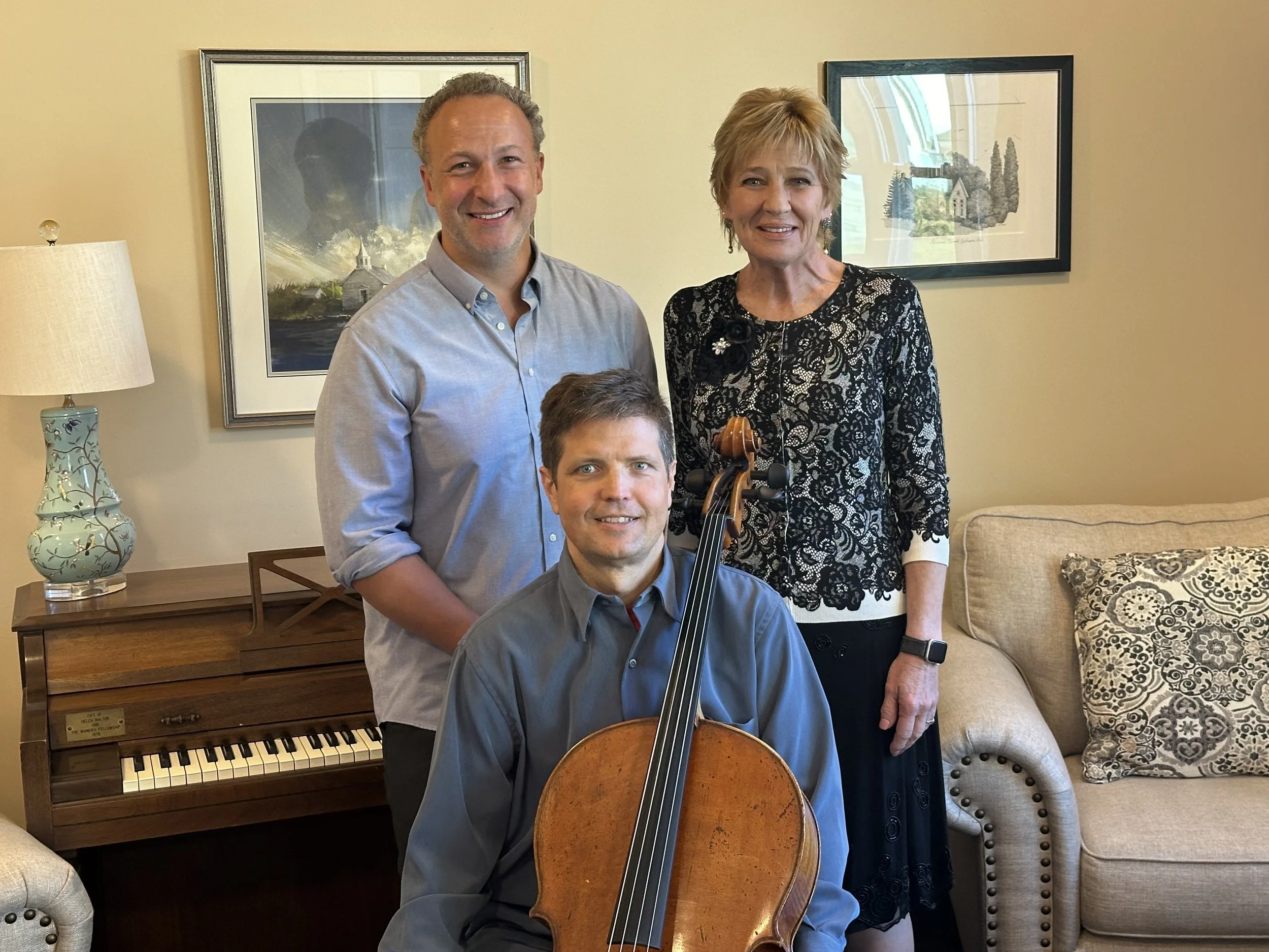 A group of four people, three men and one woman, smiling indoors. One man seated with a cello, another standing behind him, and a woman standing beside them. A piano and framed artwork are visible in the background.