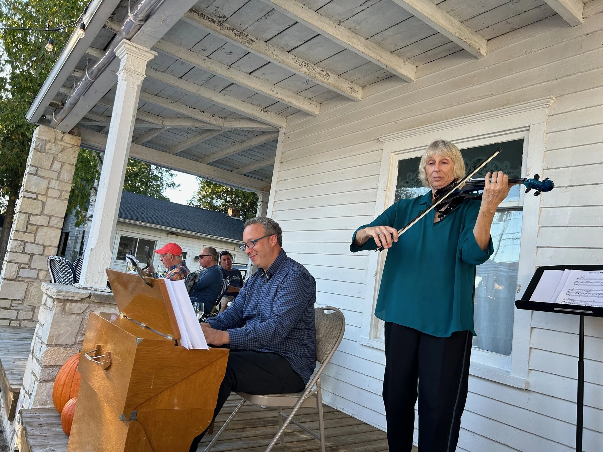 A woman playing the violin during an outdoor gathering on a porch, with several people sitting nearby, a small pumpkin on a table, and autumn decorations.