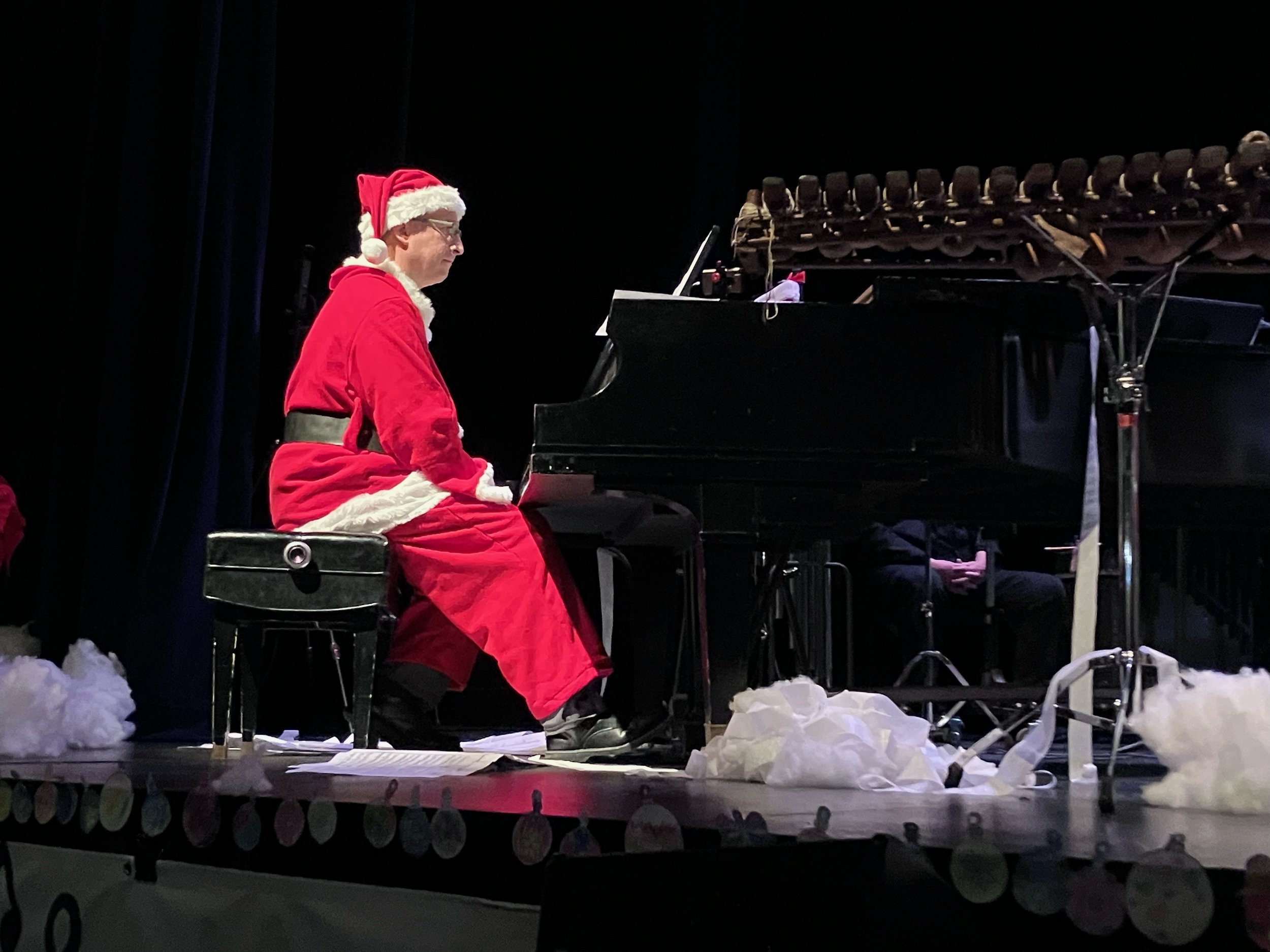 Person dressed as Santa Claus playing a grand piano on stage, surrounded by crumpled paper and snow-like props, with a dark background.