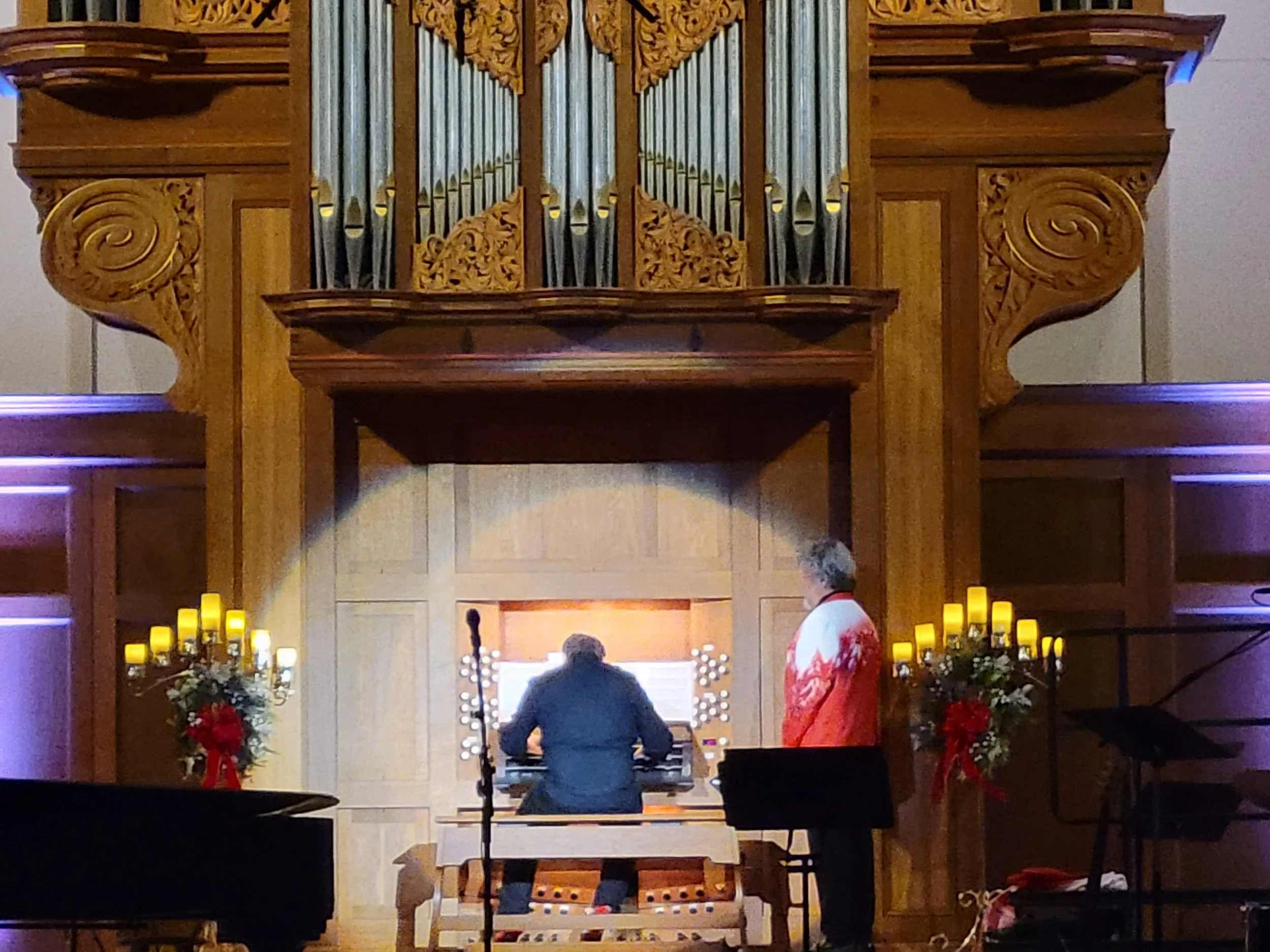 Inside a church or chapel, a person in a tuxedo is playing the piano while another person in a red and white outfit stands nearby. The area is decorated with candles and floral arrangements, likely for a special event or ceremony.