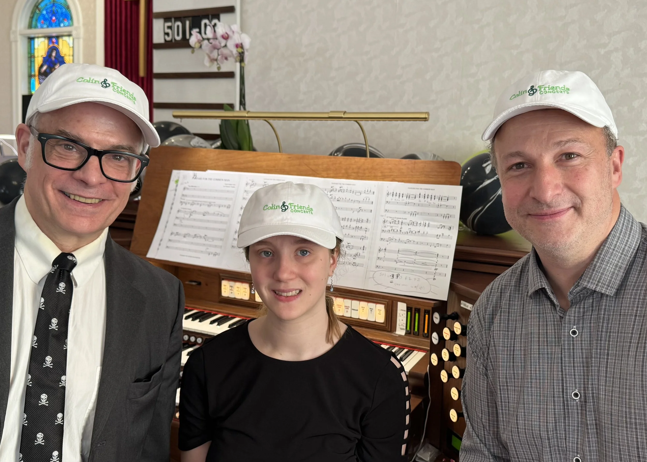 Three adults and one young girl posing together indoors, in front of a music organ with sheet music and controls, all wearing white hats with green embroidery that says 'Colin & Friends CONCERTS', smiling at the camera.