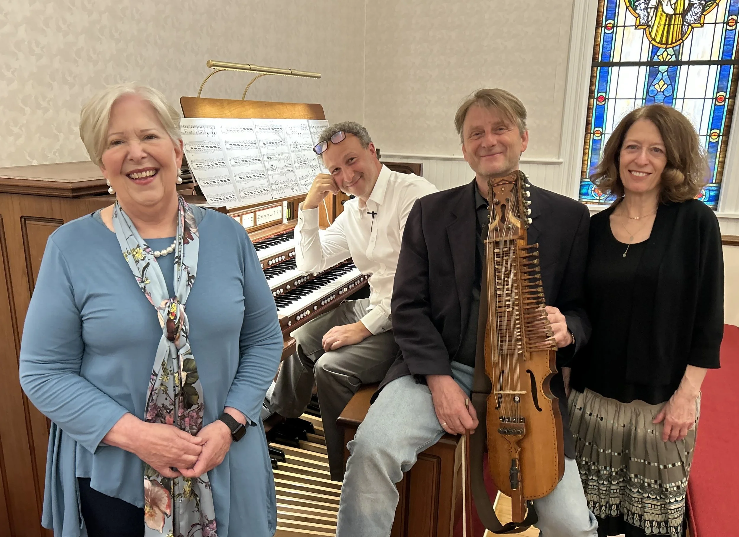 Four musicians, including a woman in a blue dress, a man with glasses playing the organ, a man holding a string instrument, and a woman with curly hair, standing inside a church with stained glass windows.
