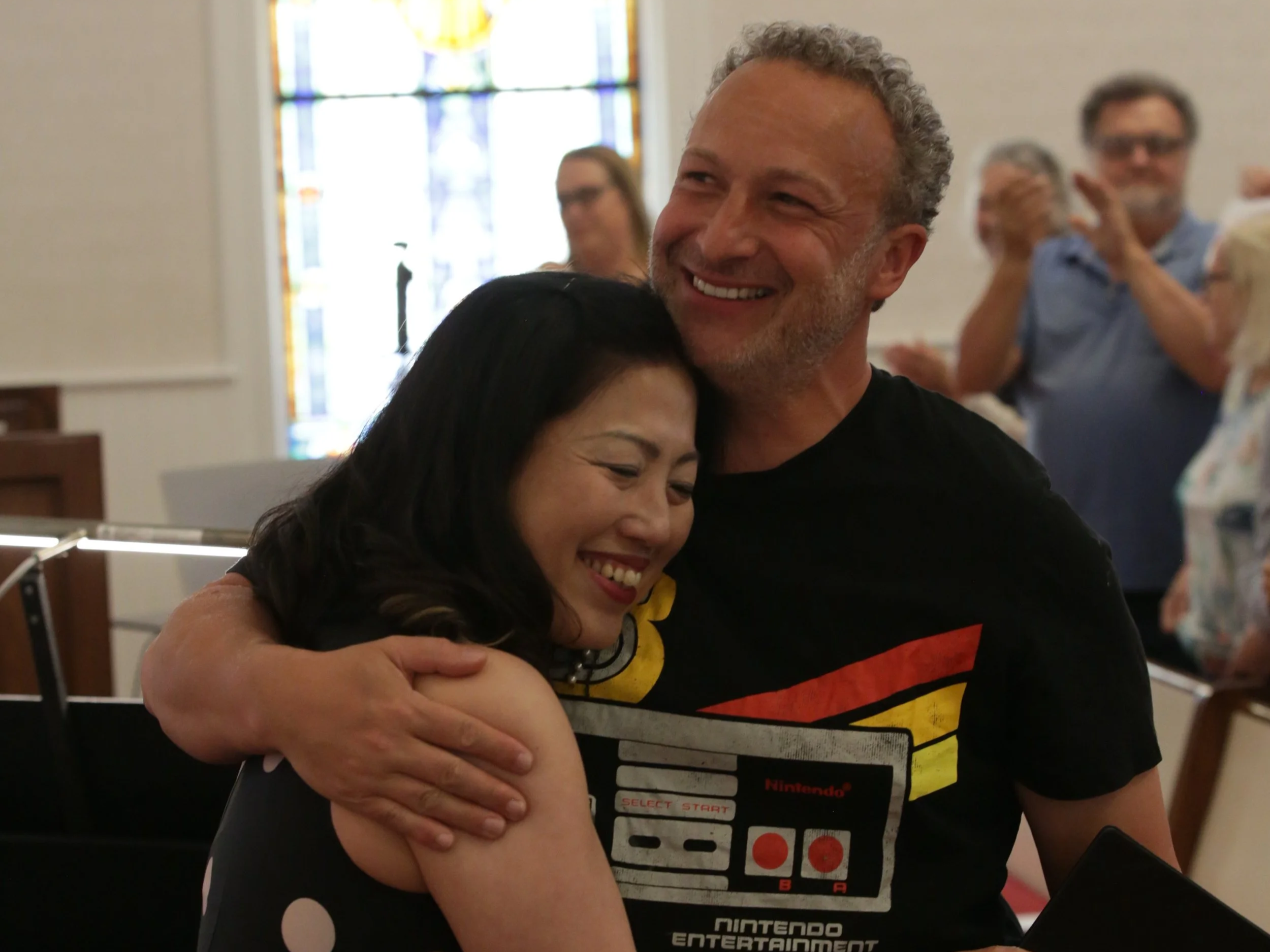 A man and woman hugging each other with smiles at a gathering or event. The man is wearing a black T-shirt with a retro Nintendo Entertainment System graphic. The background shows blurred people and a stained-glass window.