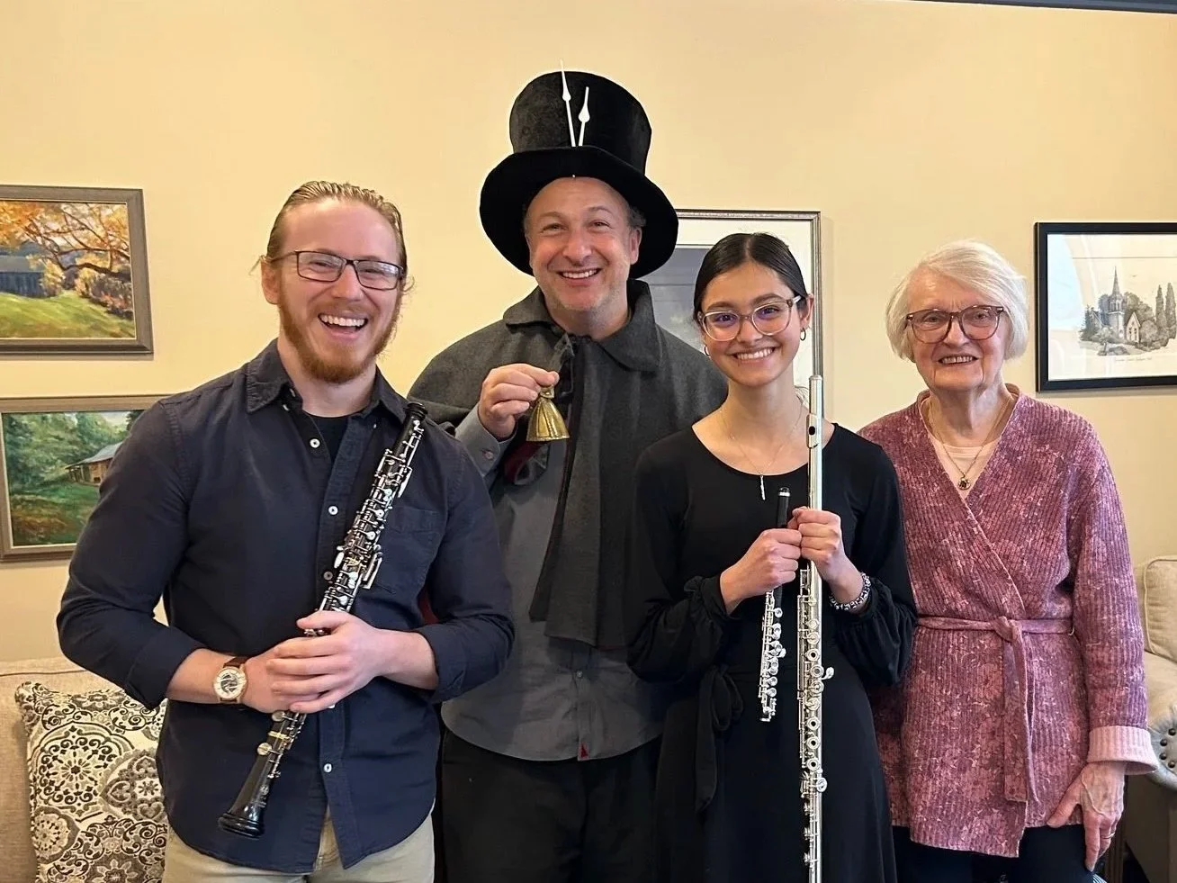 Group of five people smiling indoors, two women and three men, one man wearing a tall black hat, holding musical instruments, in front of a beige wall with framed pictures.