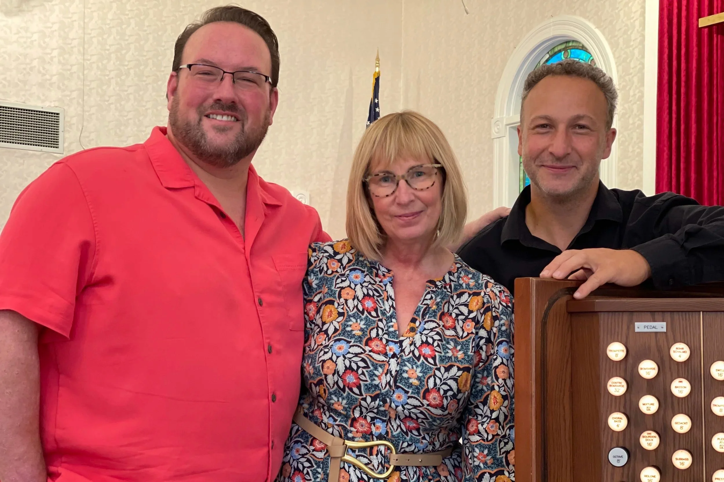 Three smiling people, two men and one woman, posing indoors with a red curtain, stained glass window, and a flag in the background. One man has glasses and a beard, wearing a red shirt; the woman is wearing glasses and a floral top, and the other man has curly hair and a black shirt.