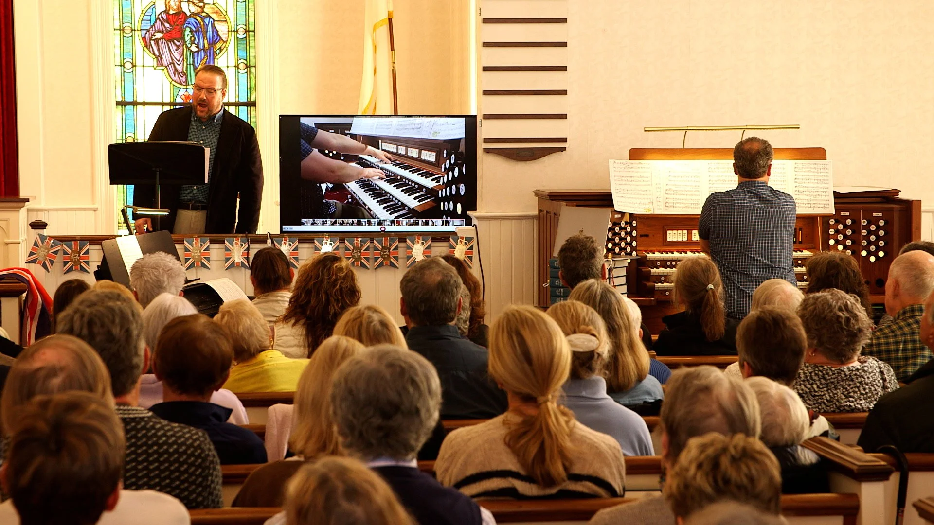 A man singing at a podium during a church service with an audience, a stained glass window, a large electronic organ, and a TV displaying a close-up of the organ.