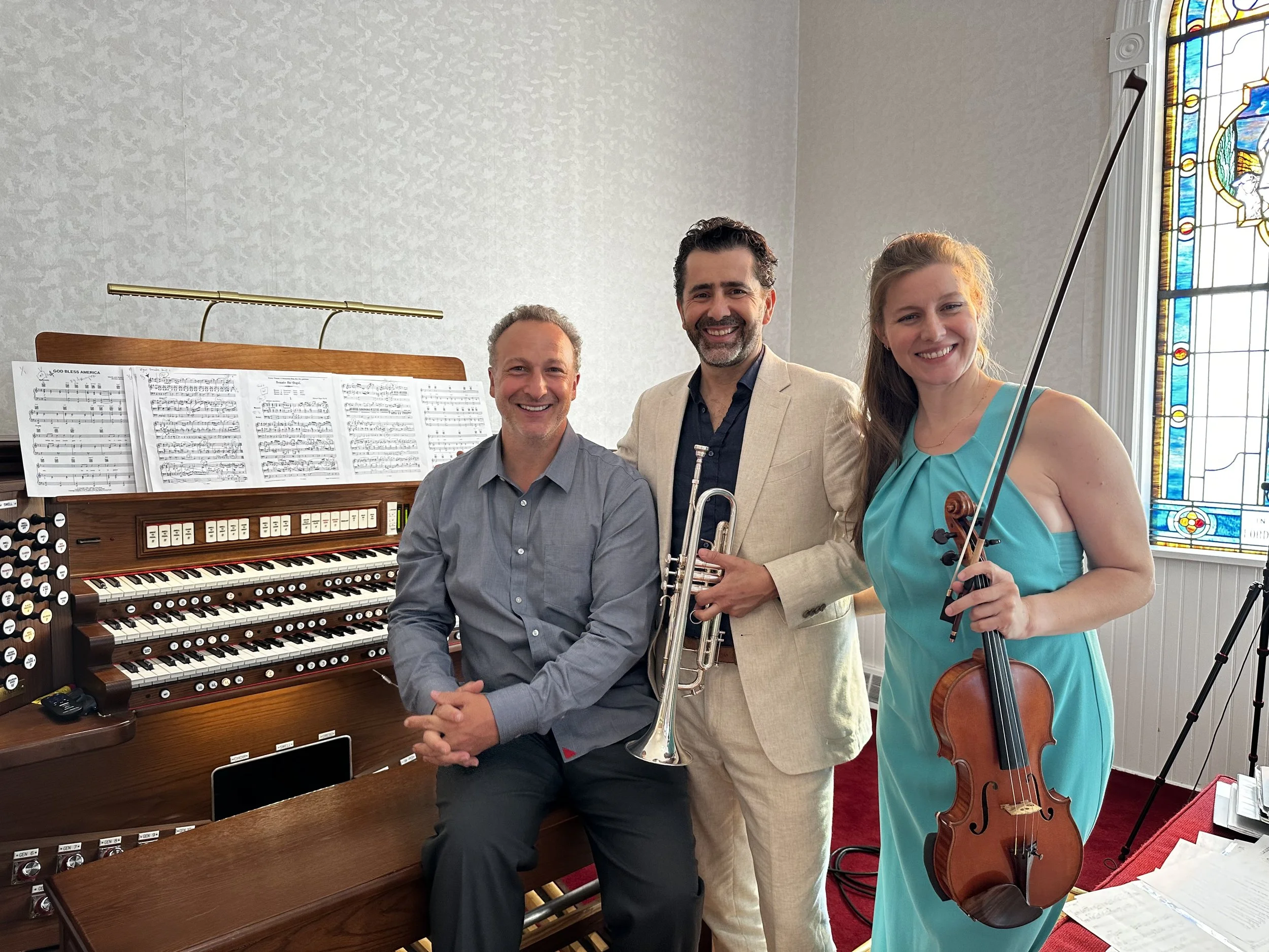 Three musicians smiling in a church with a stained glass window. One man is sitting at a Hammond organ, another man is standing with a trumpet, and a woman is standing with a violin.