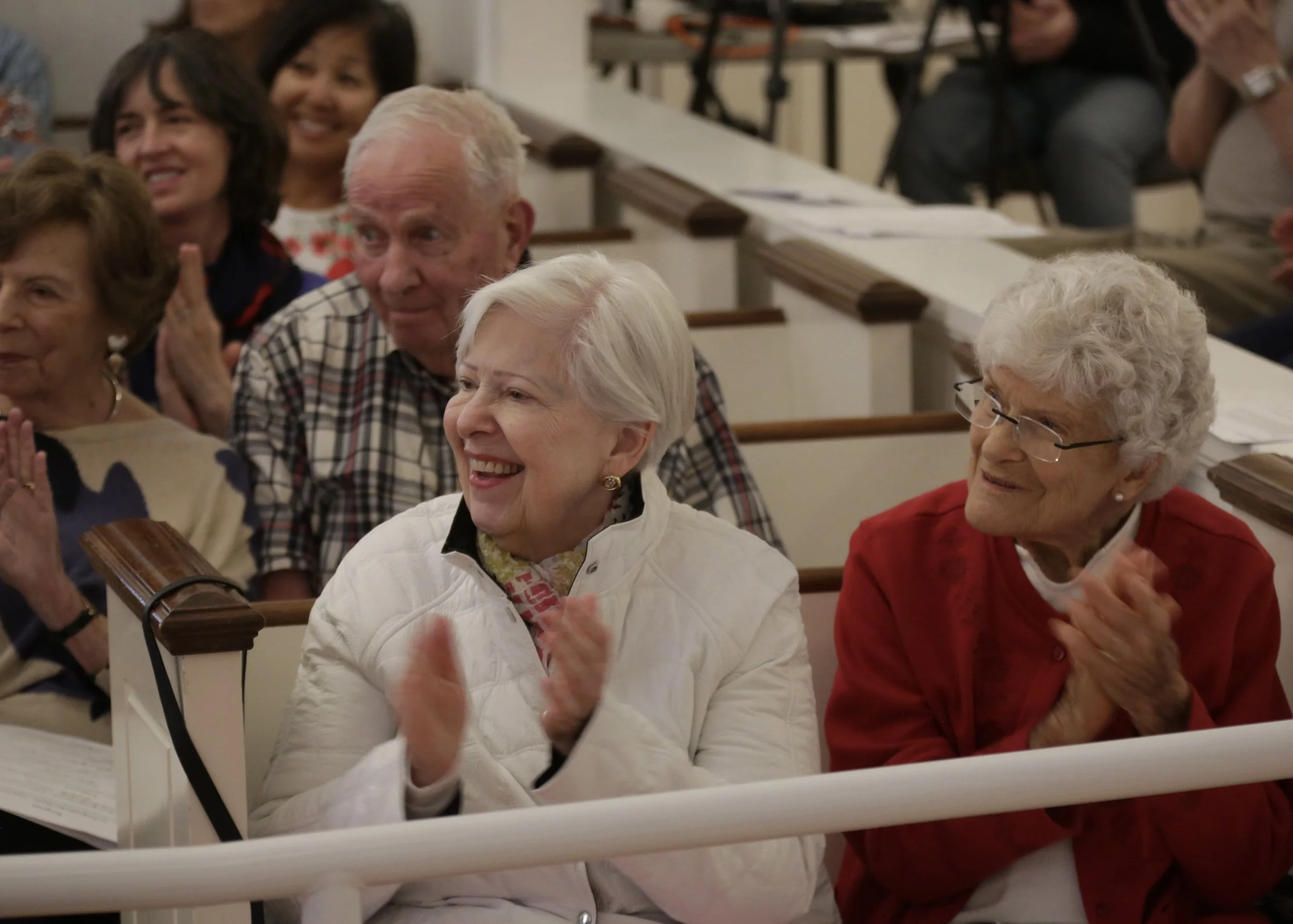A group of elderly people sitting in wooden pews, smiling and clapping, attending an event in a church or hall.