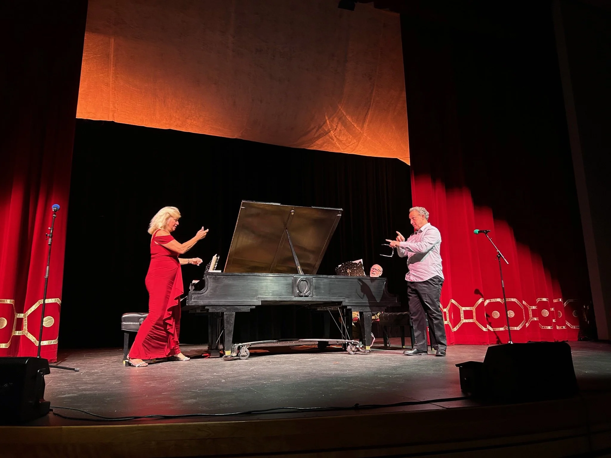 Two women and a man performing on stage with a piano in a theater with red curtains, one woman in a red dress, the man and woman appearing to be singing or playing instruments.