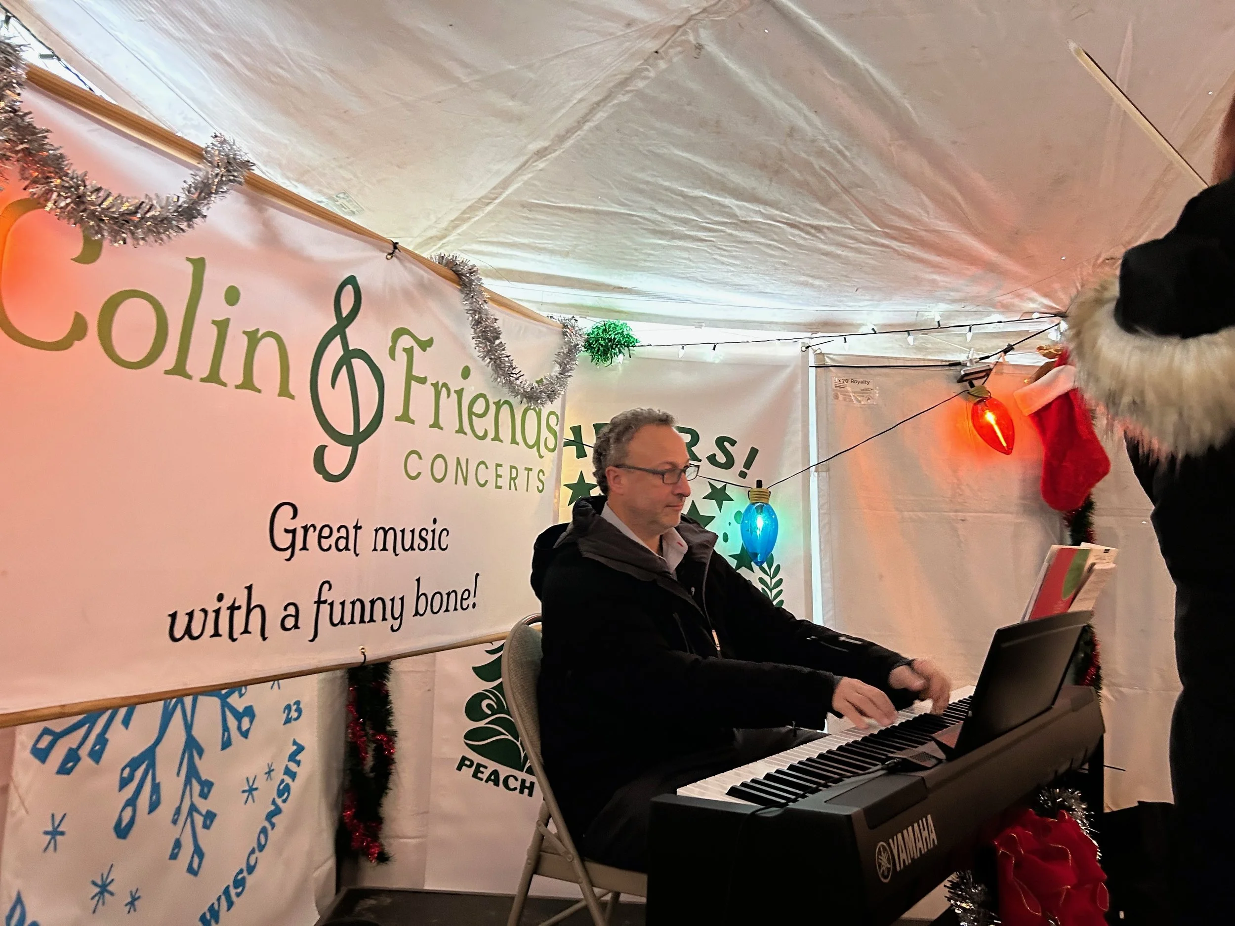 A man is playing a keyboard at a holiday concert, with a woman standing next to him, wearing a red festive outfit with white fur trim. Behind them are holiday decorations, including a banner that reads 'Holiday Friends Concerts' and Christmas tinsel.