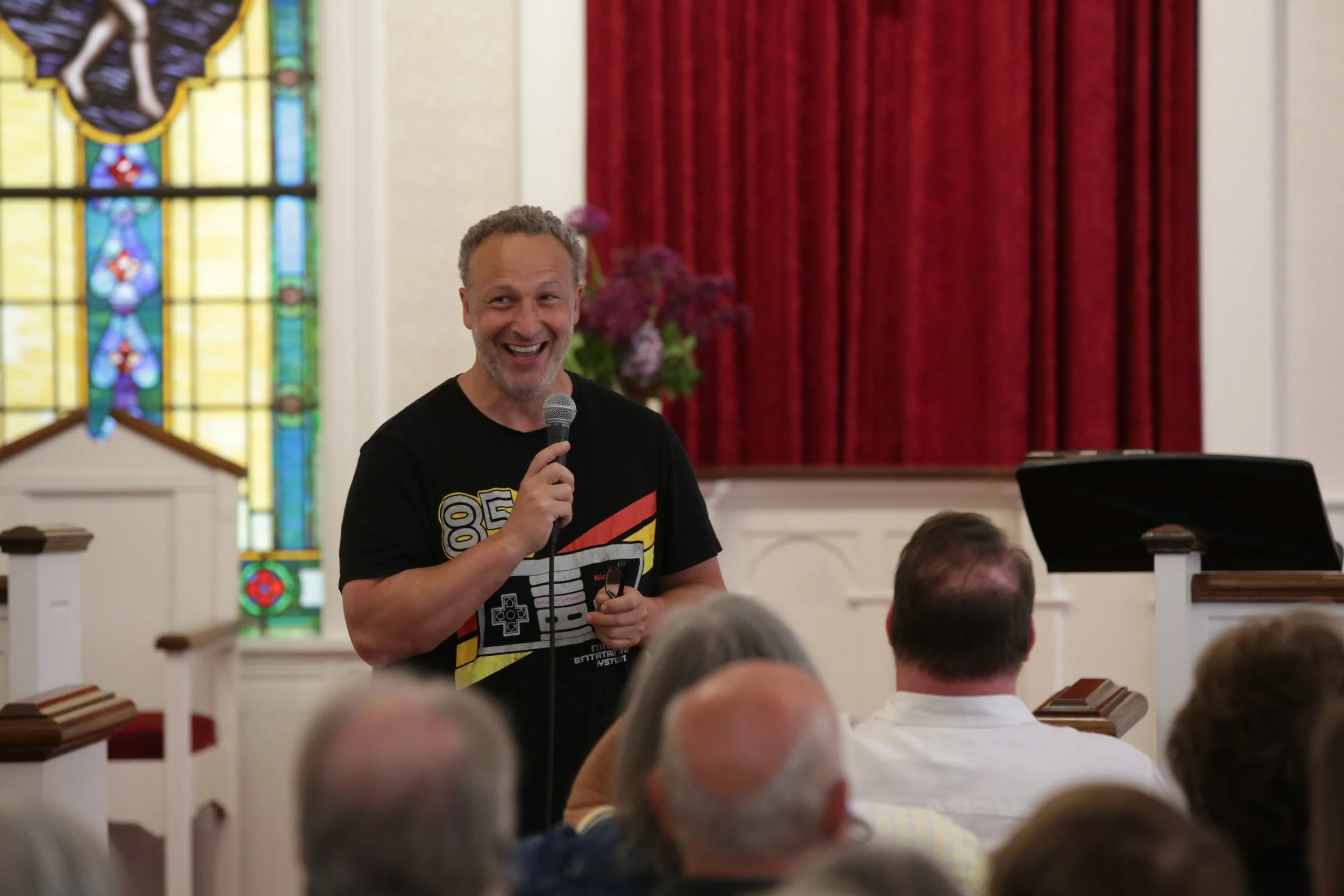 A man is smiling and speaking into a microphone during a presentation or speech inside a church or hall, with stained glass windows and red curtains in the background. Audience members are seated and visible in the foreground.
