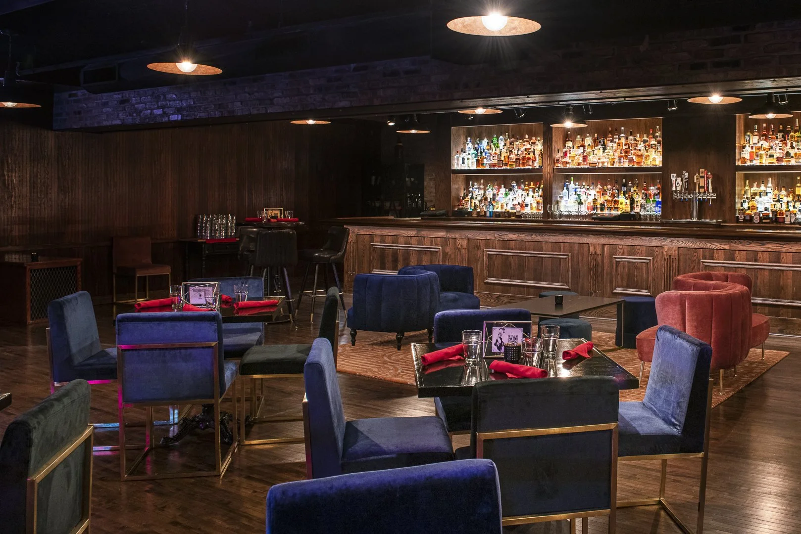 Inside a dimly lit bar with a variety of colorful bottles displayed on shelves behind a wooden counter, surrounded by modern, plush chairs and tables set with glasses and red napkins.