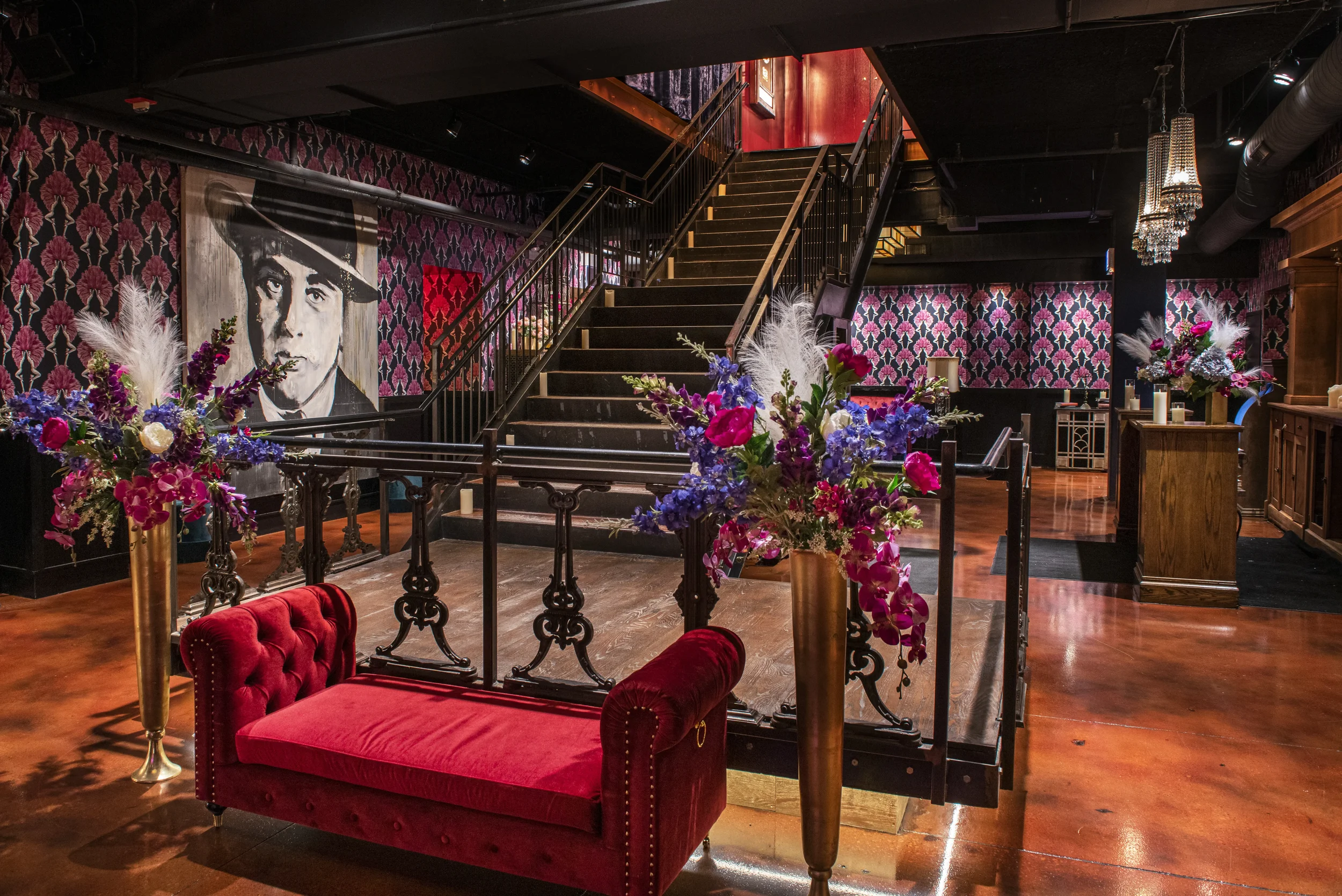 Interior of a stylish venue with a red velvet sofa, floral arrangements in gold vases, a large black and white painting of a man, a staircase, patterned wallpaper, and chandeliers.
