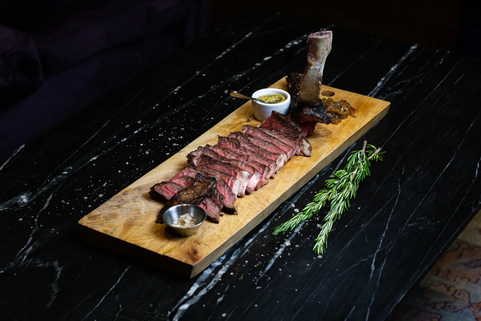 Sliced medium-rare steak with rib bone, green sauce in a cup, grilled vegetables on a wooden serving board, rosemary sprig on a dark stone table.