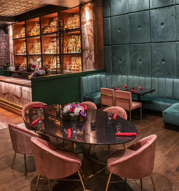 Elegant restaurant or lounge with pink velvet chairs around a black marble table set with red cloth napkins, floral centerpieces, and small glasses, featuring a large bar with illuminated shelves filled with bottles in the background and a green upho