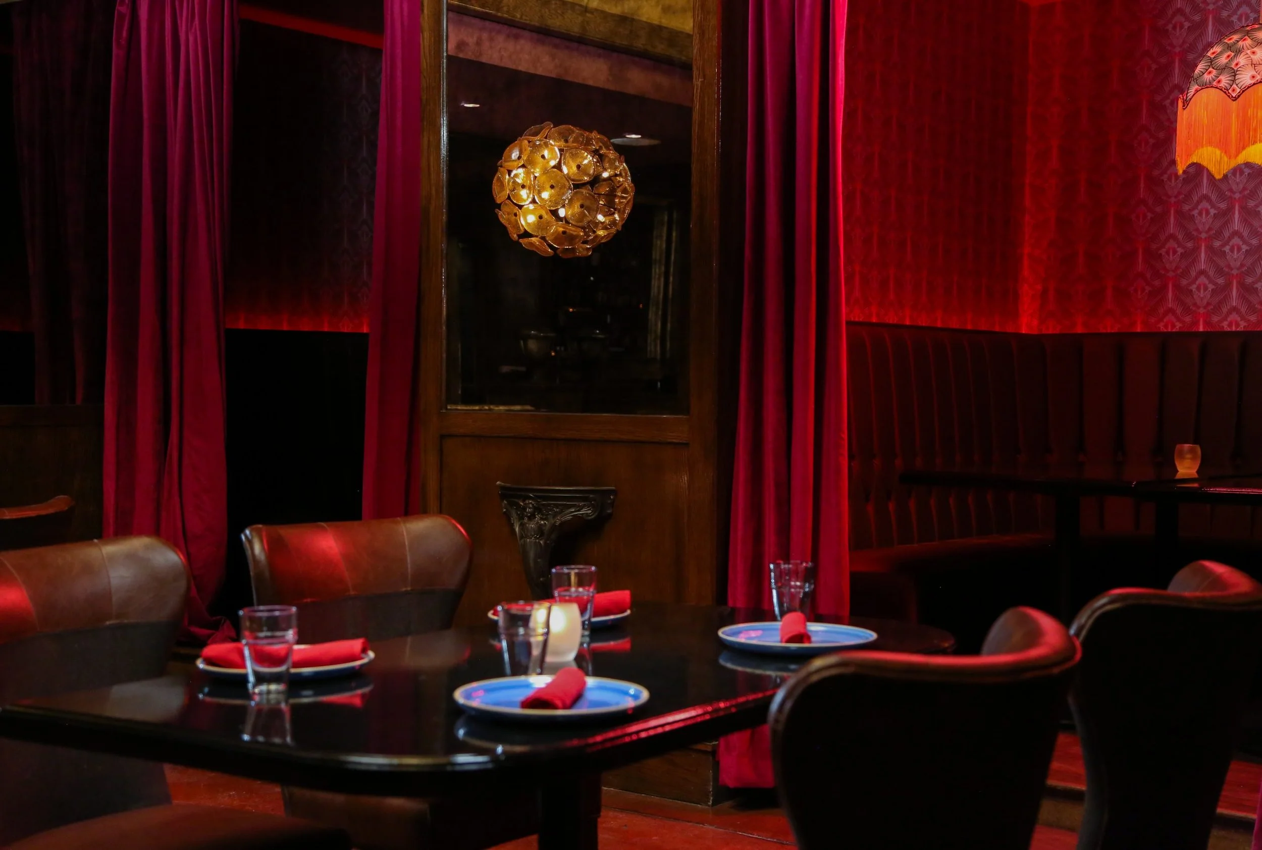 Elegant restaurant interior with dark wooden furniture, red curtains, and red wall accents. A round chandelier hangs from the ceiling, and the table is set with blue plates, red napkins, glasses, and candles.
