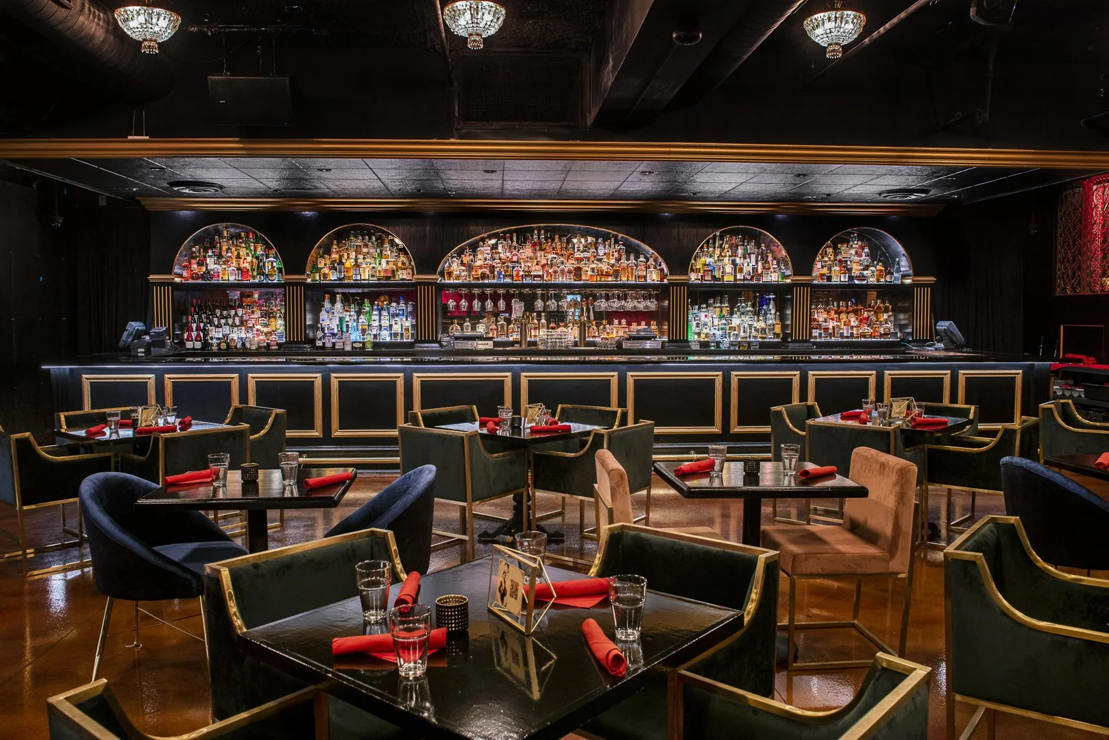 Empty upscale bar and restaurant with black and gold decor, tables set with glasses, red napkins, and small centerpieces, and a fully stocked bar in the background.