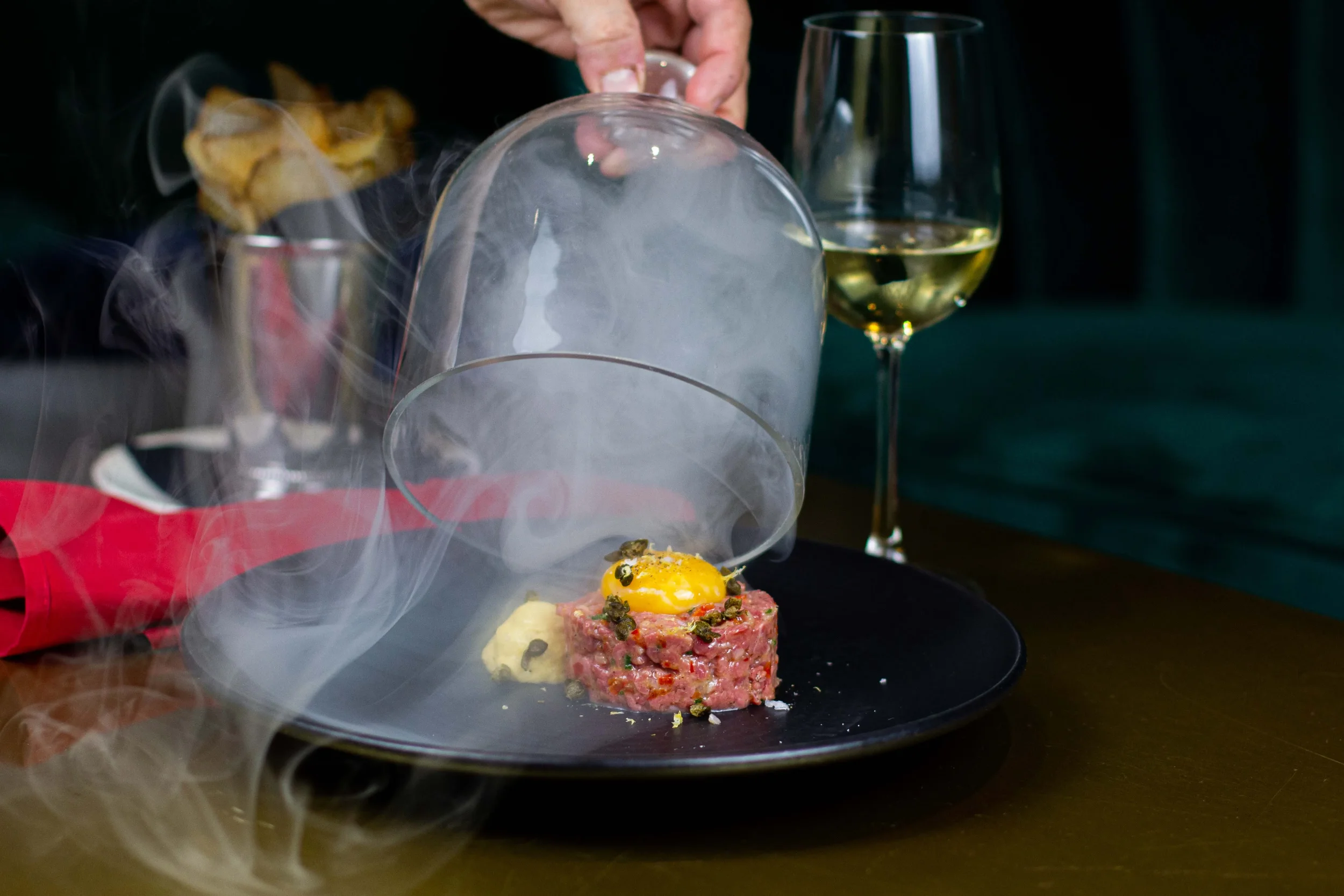 A dish of steak tartare topped with a raw egg yolk, with smoke or vapor rising from it, served on a black plate next to a glass of white wine.