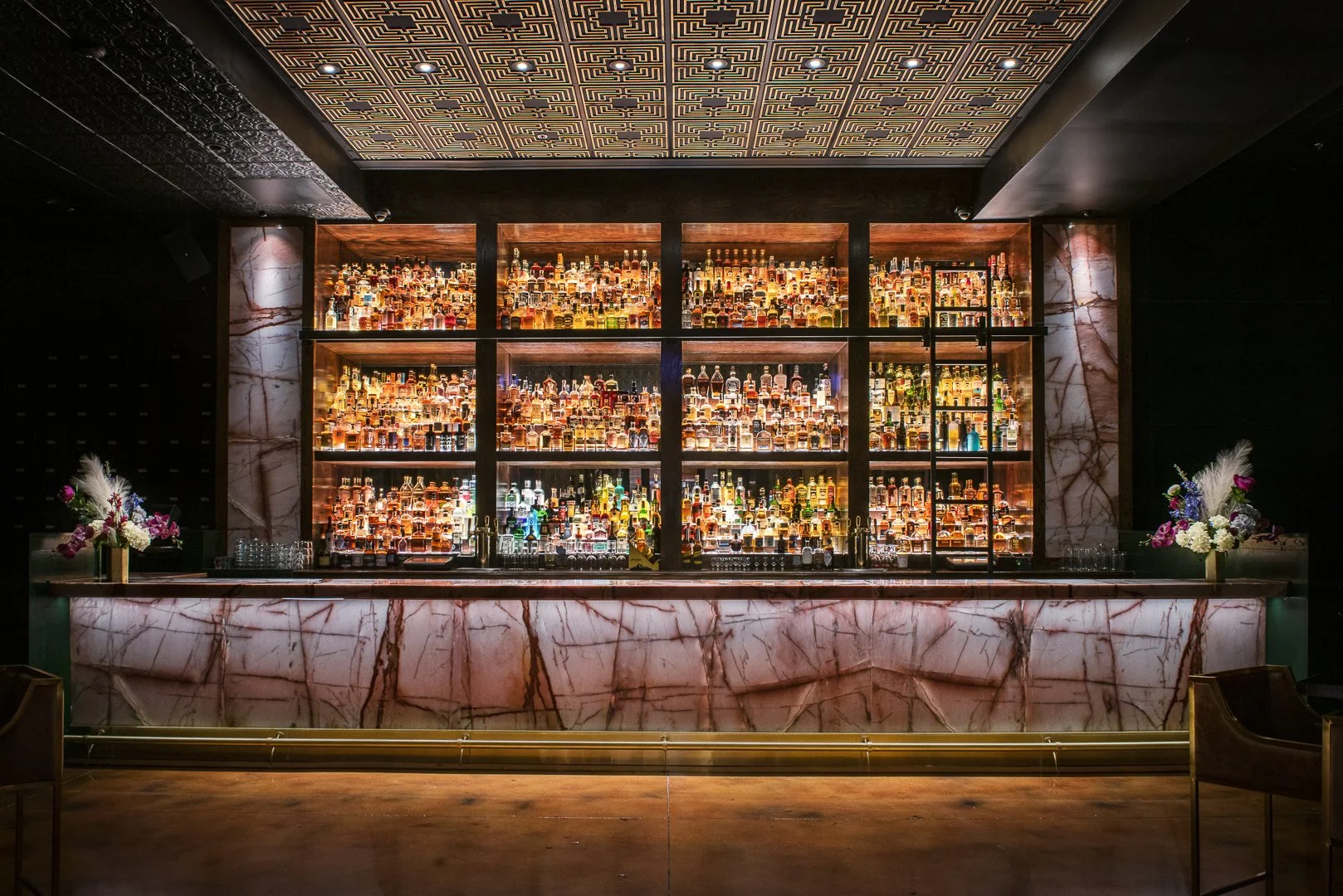Luxurious bar with illuminated shelves filled with bottles of liquor, marble countertops, and floral arrangements on either side.
